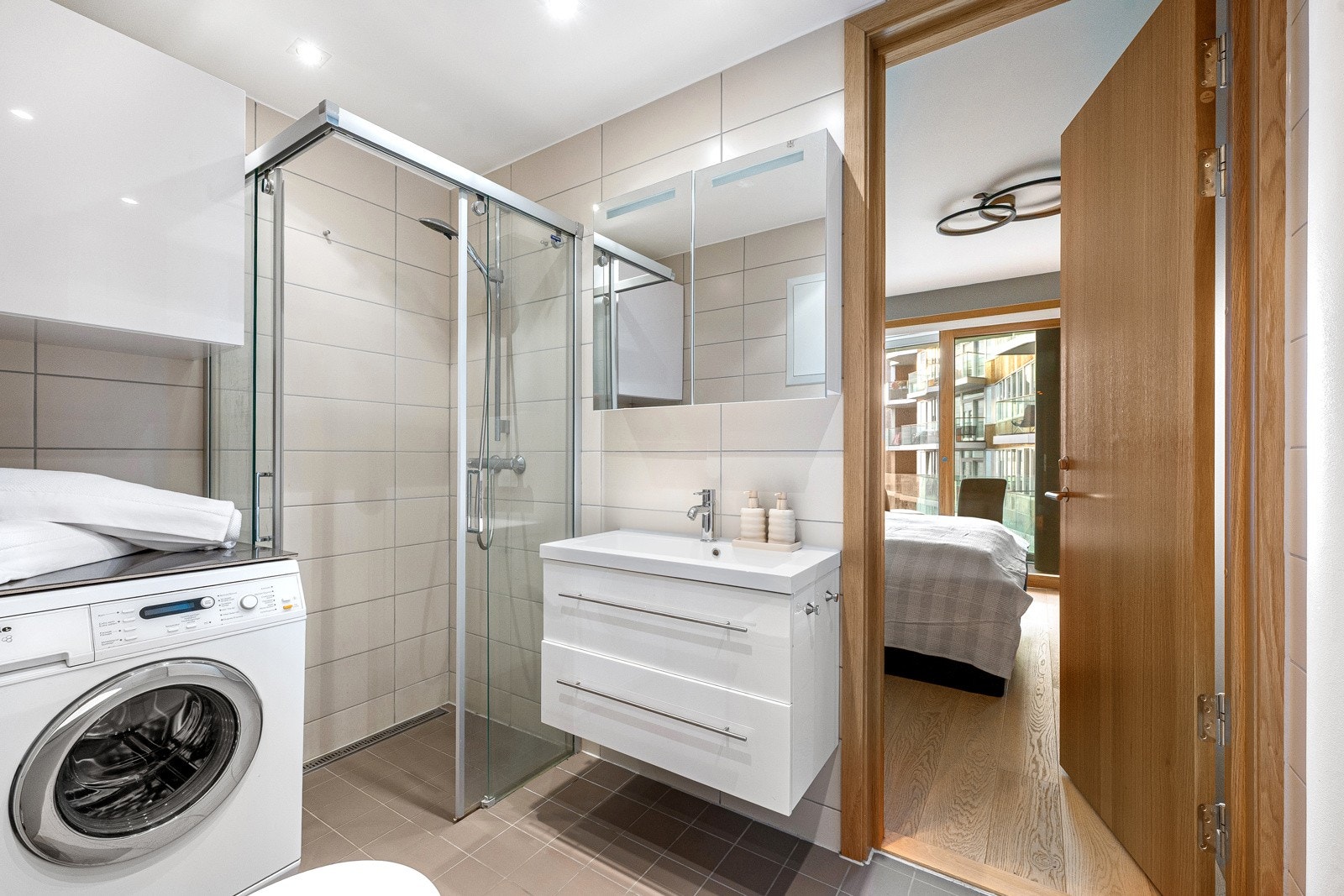 Stilrent bad med vegghengt wc, opplegg til vaskemaskin og tørketrommel, dusjhjørne med glassvegger og baderomsinnredning med oppbevaringsplass i skuffer og skap Galleribilde