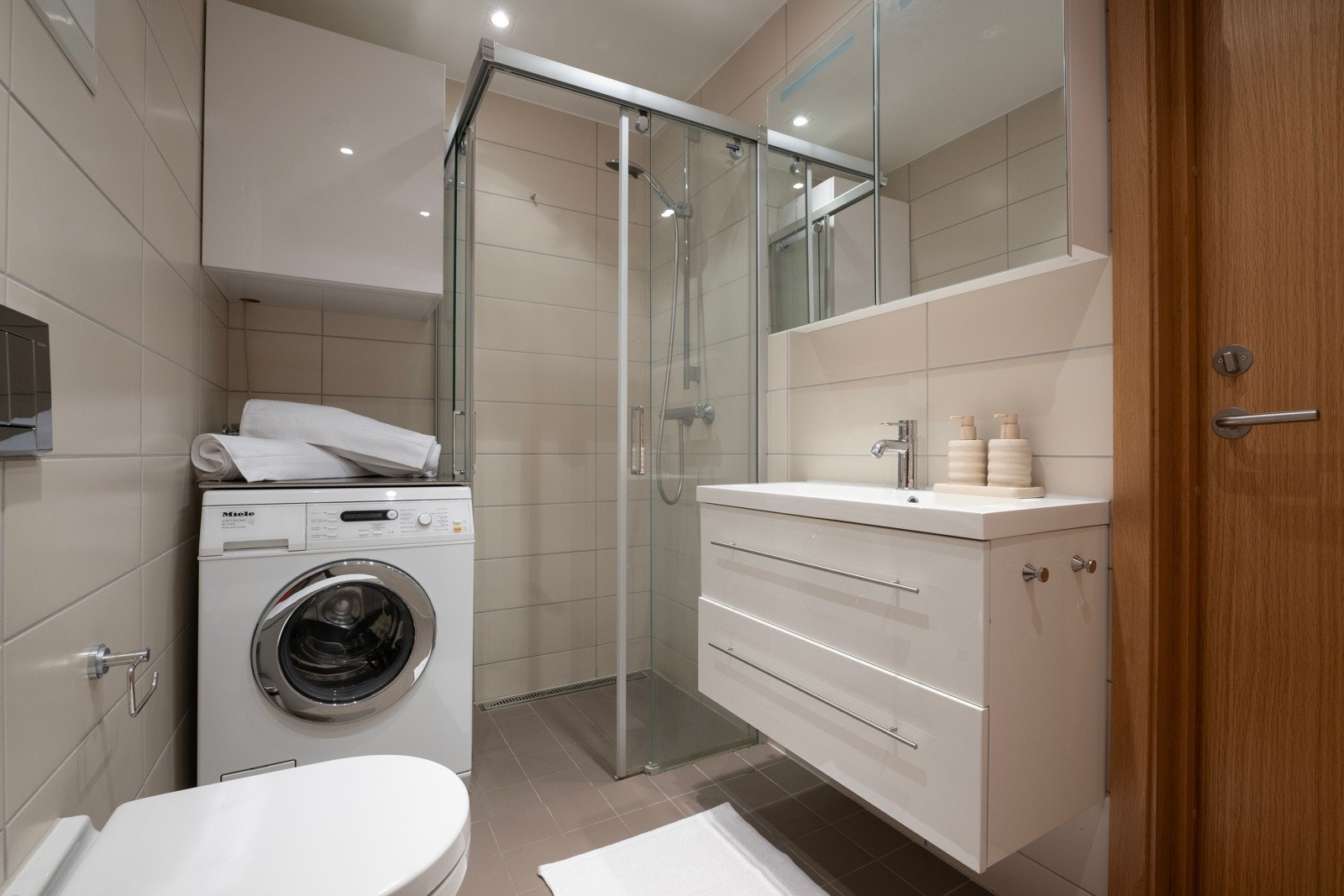Stilrent bad med vegghengt wc, opplegg til vaskemaskin og tørketrommel, dusjhjørne med glassvegger og baderomsinnredning med oppbevaringsplass i skuffer og skap Galleribilde