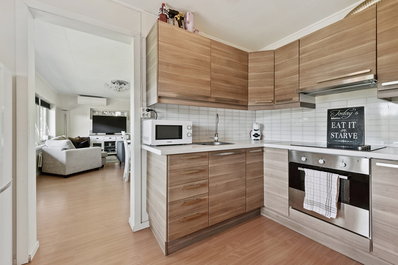 Pen kjøkkeninnredning med glatte fronter, fliser over kjøkkenbenk, integrert koketopp og stekeovn, samt åpen løsning til stue. Galleribilde