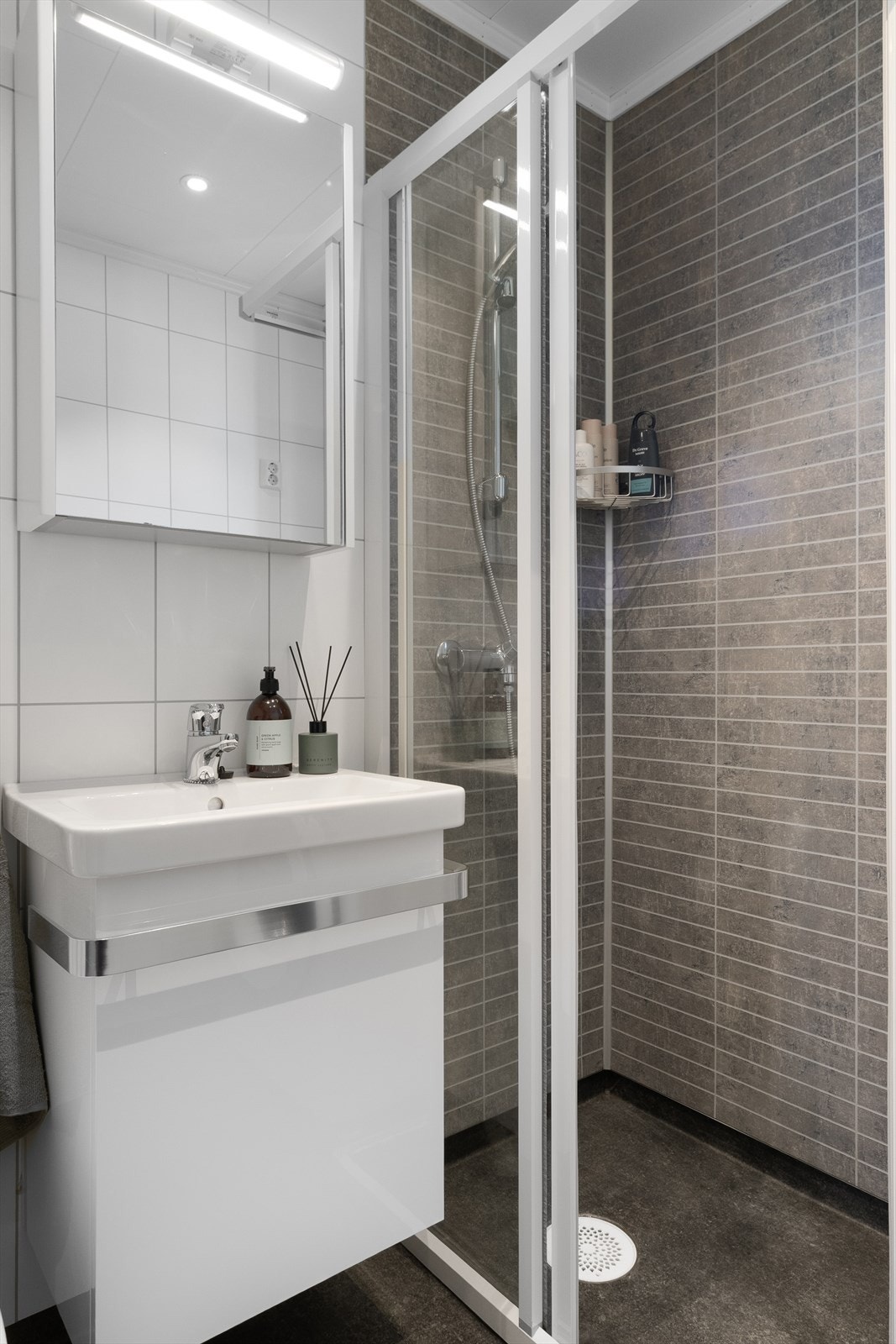 Rommet er innredet med et servantskap med servant, gulvmontert toalett og en vegghengt dusj med skjermdører i glass/aluminium. Galleribilde