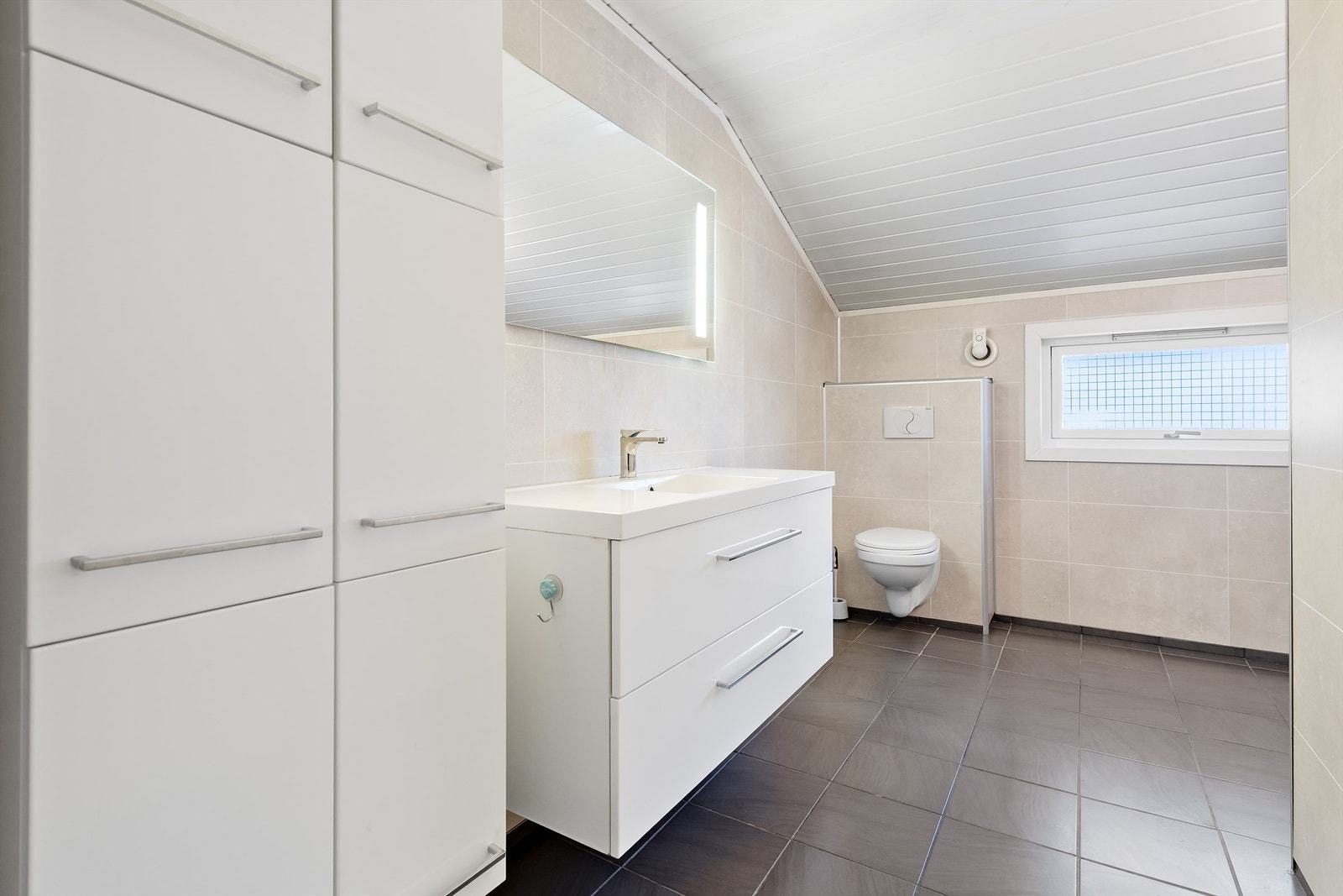 Badet i 1. etasje har flis på gulv med varmekabler, våtromsplater på veggene, hvit innredning og vegghengt toalett. Galleribilde