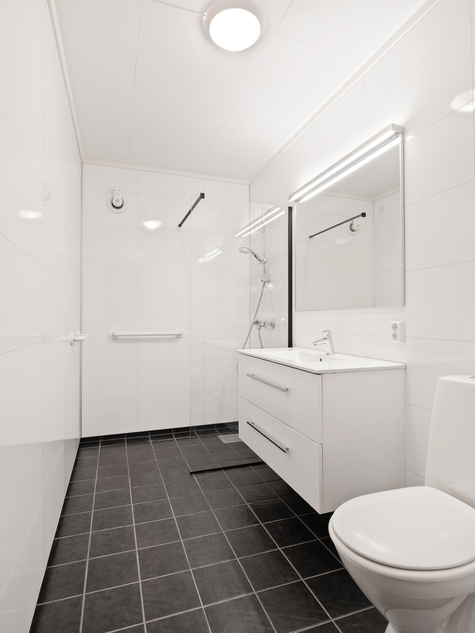 Det er lyse baderomsplater på vegger og mørke fliser med varmekabler på gulvet Galleribilde