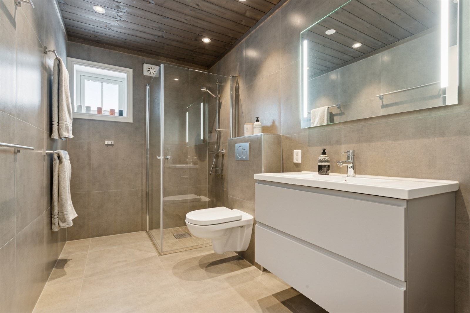 Baderommet er delikat innredet med store, grå fliser på gulv og vegger. I gulvet har man også varmekabler for ekstra komfort. Badet er ellers utstyrt med dusjhjørne, vegghengt toalett og baderomsinnredning med god lagringsplass og nedfelt servant. Galleribilde