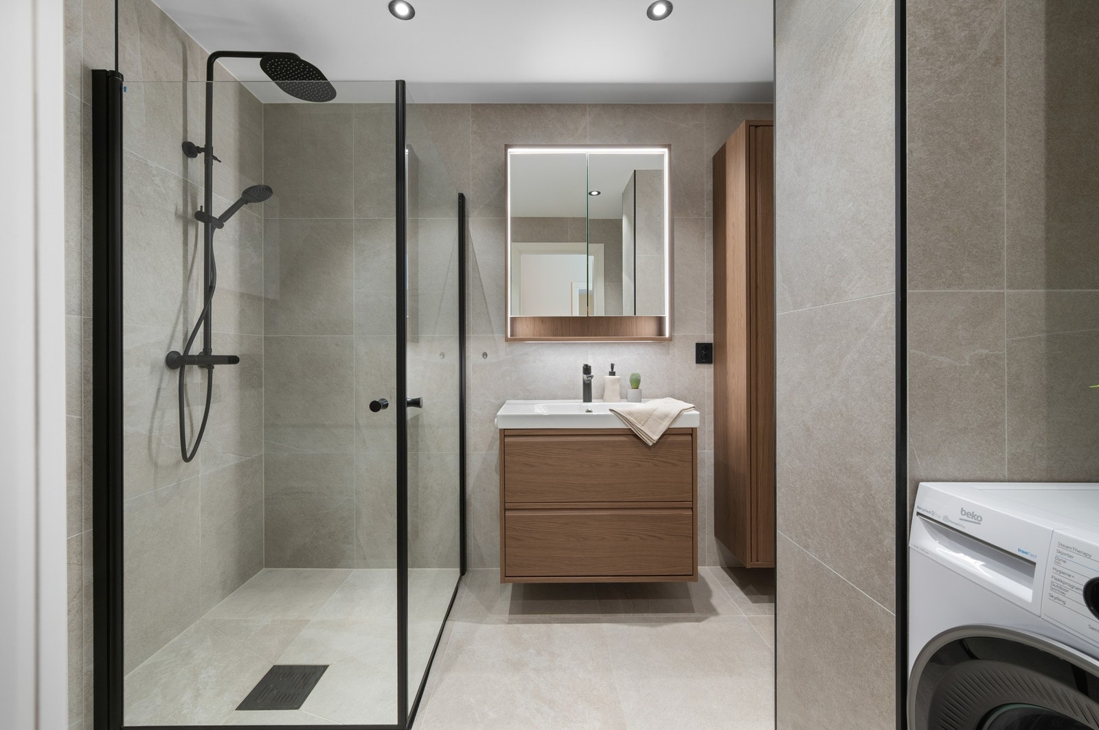 Badet inneholder vegghengt toalett, helstøpt servant, skuffer under servant med slette fronter, dusjdører i klart glass og opplegg for vaskemaskin. Galleribilde