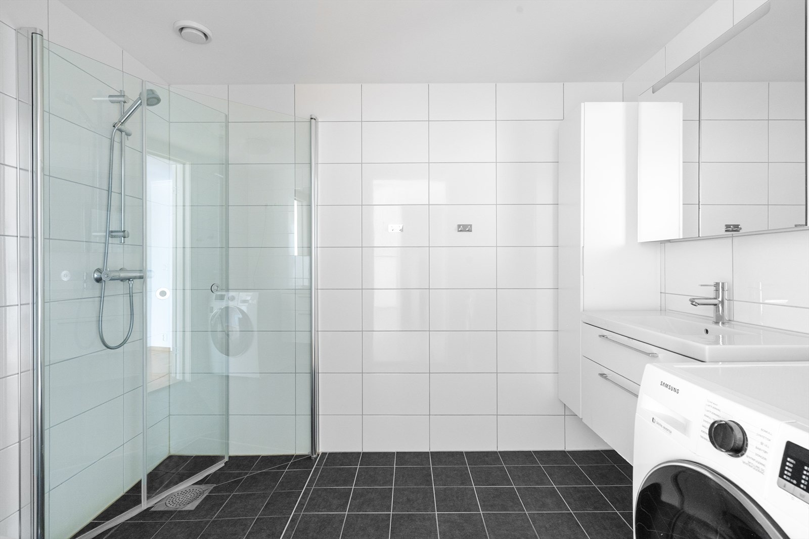 Badet, som også fungerer som vaskerom med opplegg for vaskemaskin, er helfliset med varmekabler i gulvet. Moderne innredning med skuffer og høyskap. Badet er også innredet med dusjnisje med innfellbare glassvegger og veggmontert toalett. Galleribilde