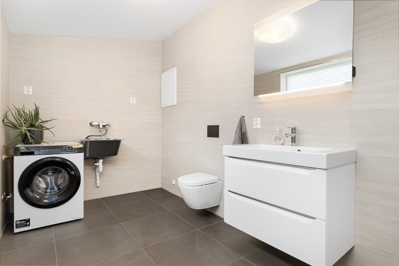 Lyst og stilrent bad/vaskerom med flis på gulv og vegger. Rommet har gulvvarme og er utstyrt med opplegg for vaskemaskin, servant, servantskap, wc, dusjkabinett og rørfordelingsskap. Galleribilde