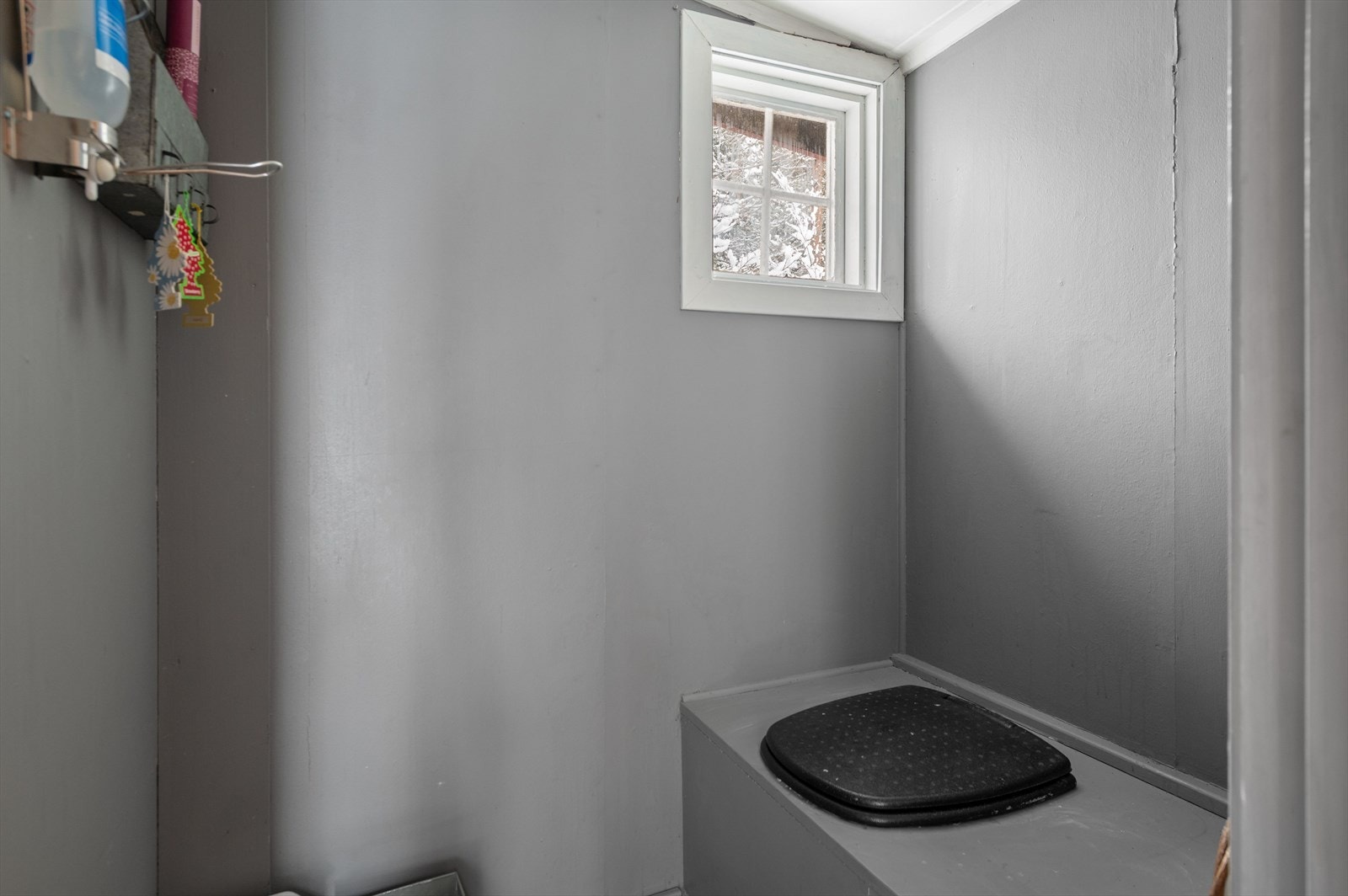 Gråmalte vegger og et lite vindu gir enkel belysning og privatliv i rommet som har tradisjonell utedo-løsning. Galleribilde