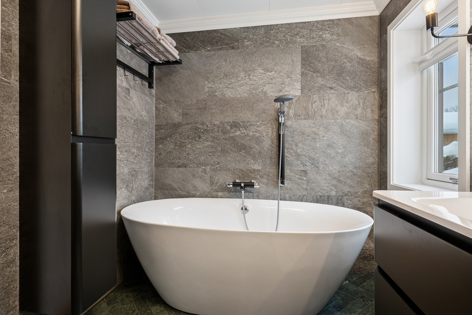 Badet har mørke lekre fliser og innredet med servant med underskap, speil med belysning, vegghengt dusj og et badekar med dusjfunksjon. Galleribilde