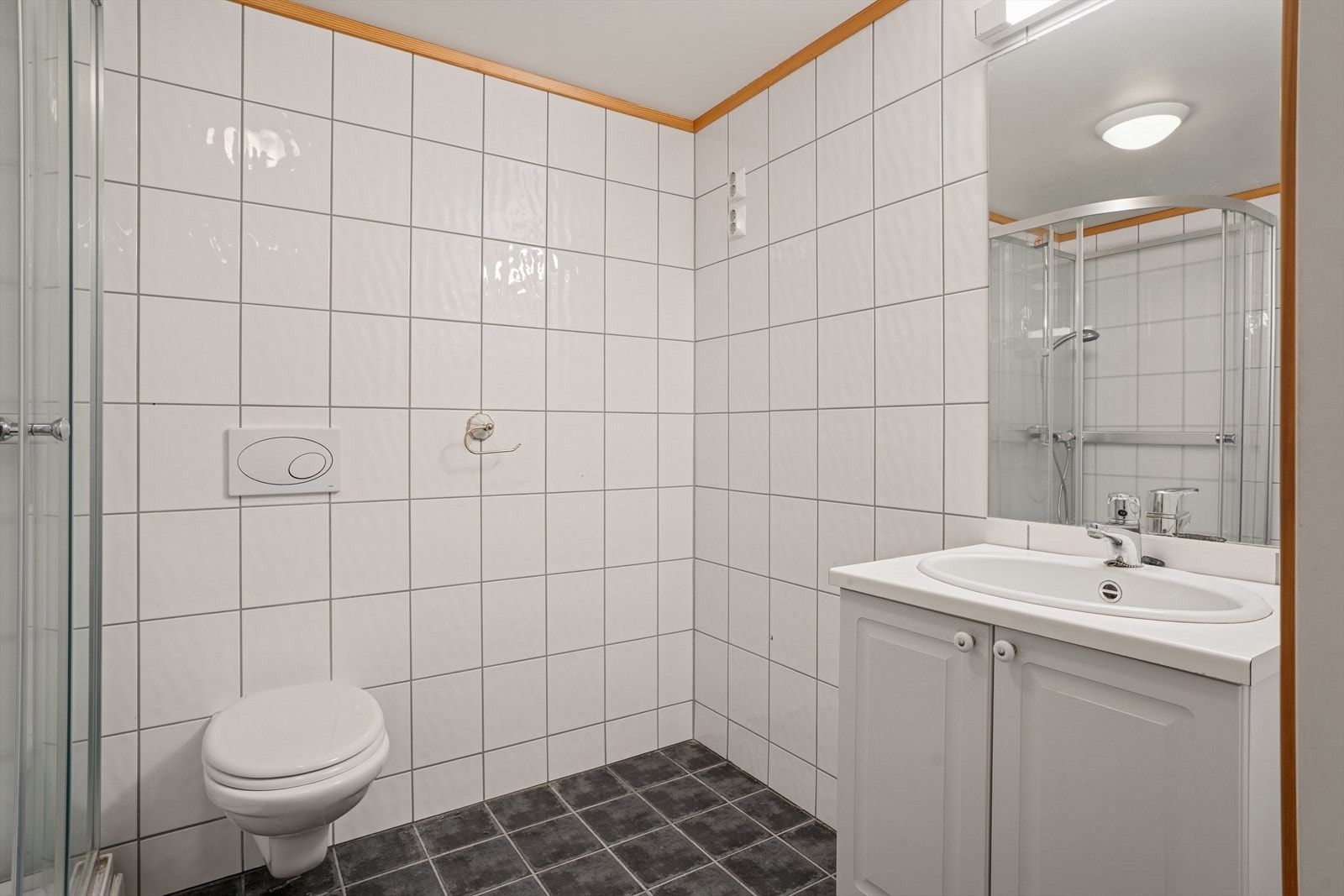 Bad med flislagte overflater på gulv og vegger. Badet er innredet med baderomsmøbel med nedfelt servant med profilerte, lyse fronter, høyskap og overhengende speil med belysning. Galleribilde