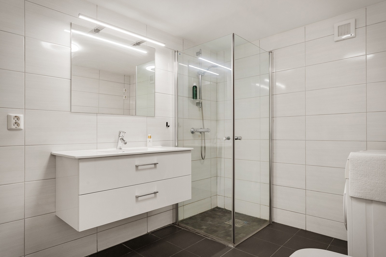 Bad innredet med vegghengt toalett, dusj med glass foldedører, servant med møblement, speil og belysning. Røropplegg for vaskemaskin. Galleribilde