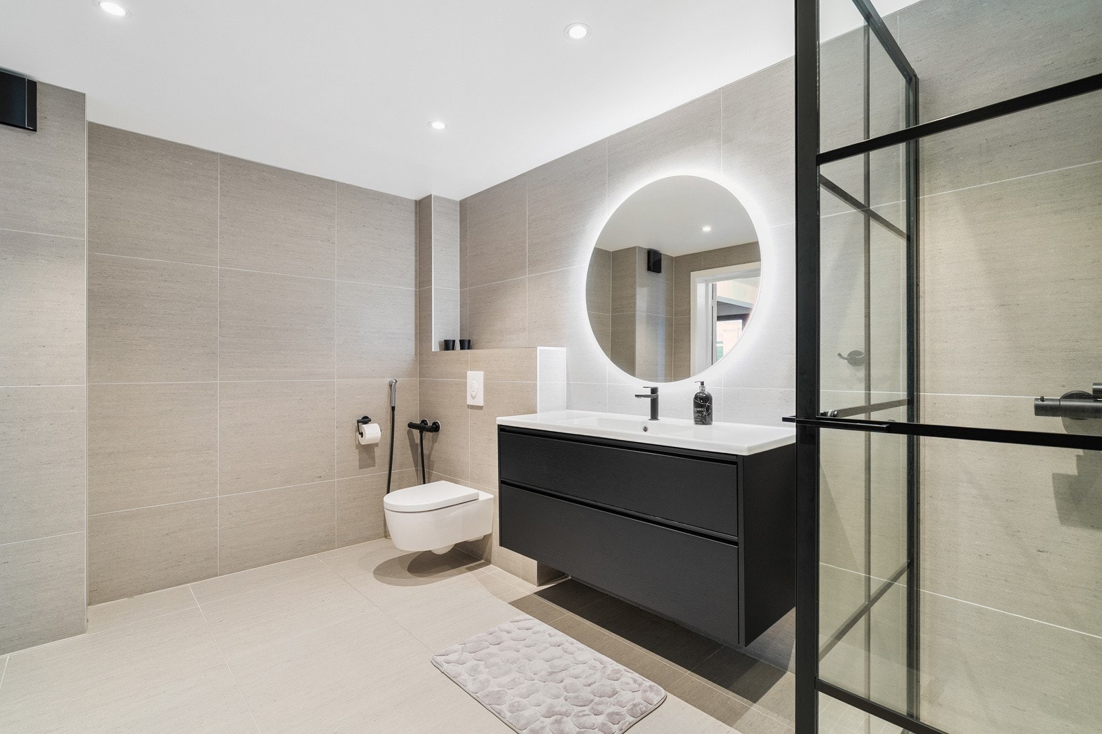 Moderne baderom med stilrene detaljer, flislagte overflater og sort innredning. Rommet er utstyrt med vegghengt toalett, dusjhjørne og servant med oppbevaringsskuffer. Galleribilde