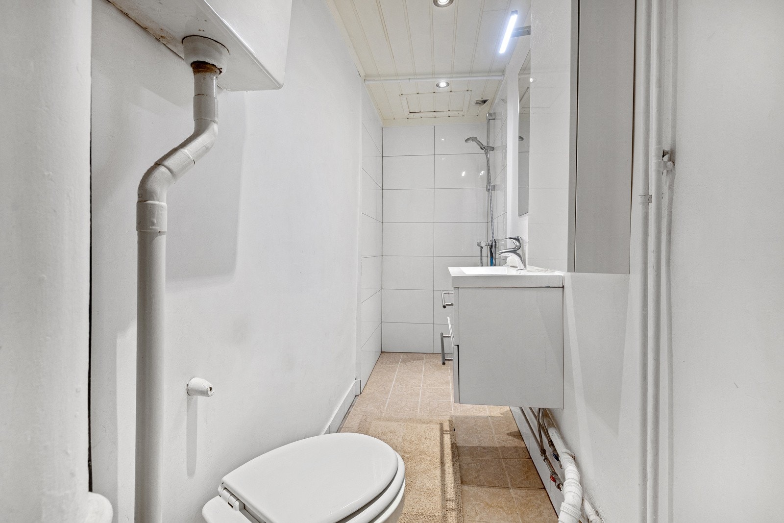 Badet har en smal utforming og er innredet med et servantskap med glatte skuffefronter, gulvstårende toalett og dusjhjørne. Galleribilde