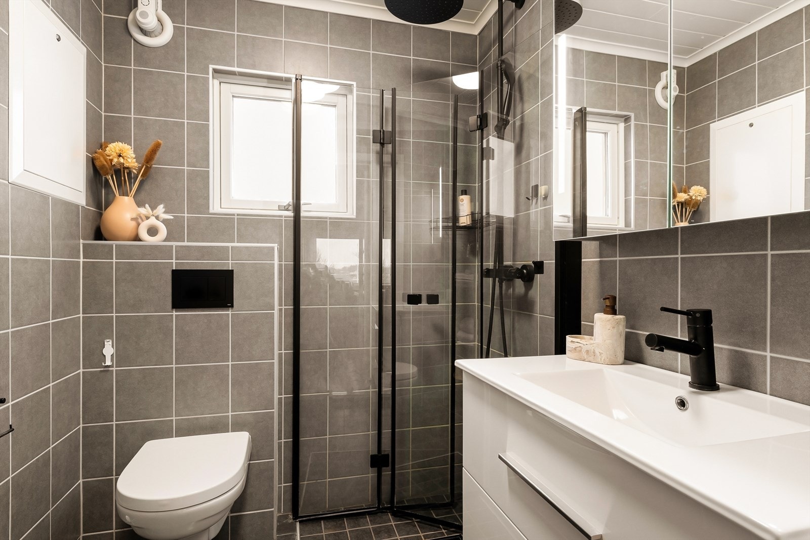 Badet har flis på gulv og vegger o malt panel i taket. Servant i skuffinnredning, speilskap, vegghengt toalett og foldbare dusjdører i glass. Galleribilde