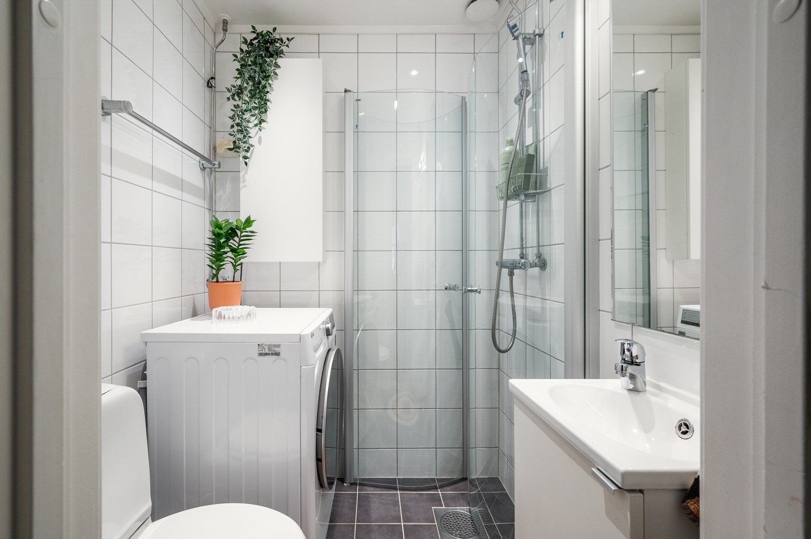 Pent og delikat flislagt baderom med dusjhjørne med svingbare glassdører, servant med underskap, vegghengt speil og toalett. Galleribilde