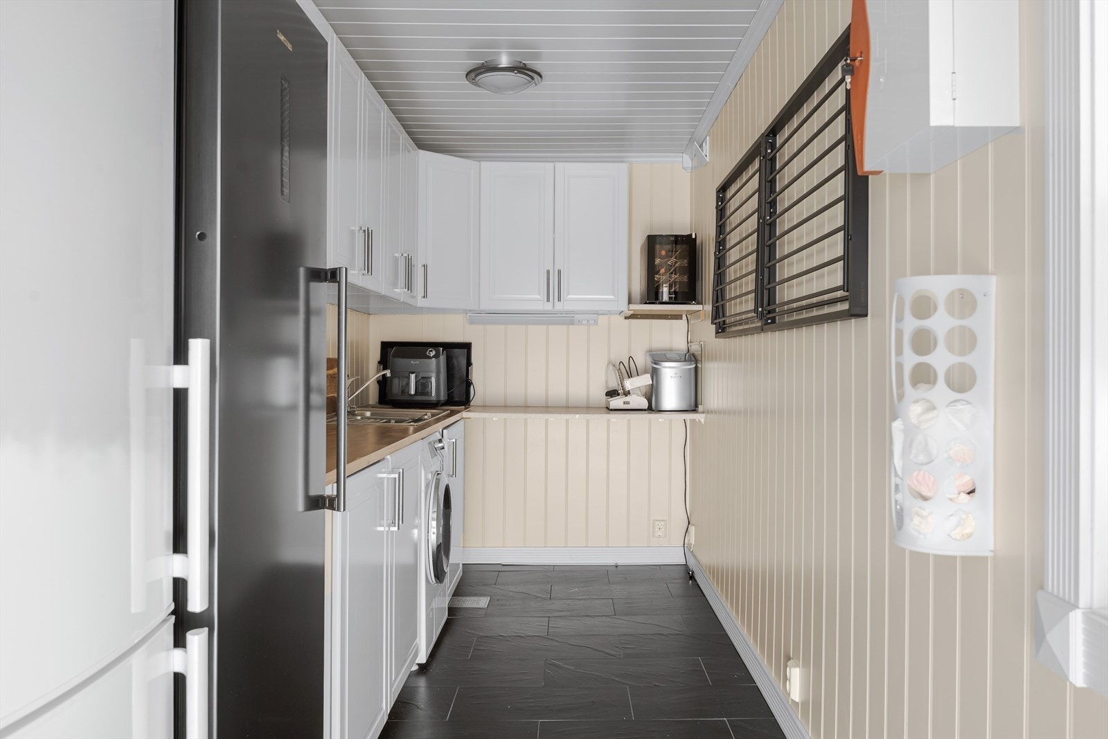 Vaskerom/grovkjøkken fra tilbyggningsårene ca. 2012/13. Lys innredning med profilerte fronter og benkeplate med to nedfelte kummer. Plass og opplegg for vaskemaskin under benkeplaten. 2 vegghengte tørkestativ. Galleribilde