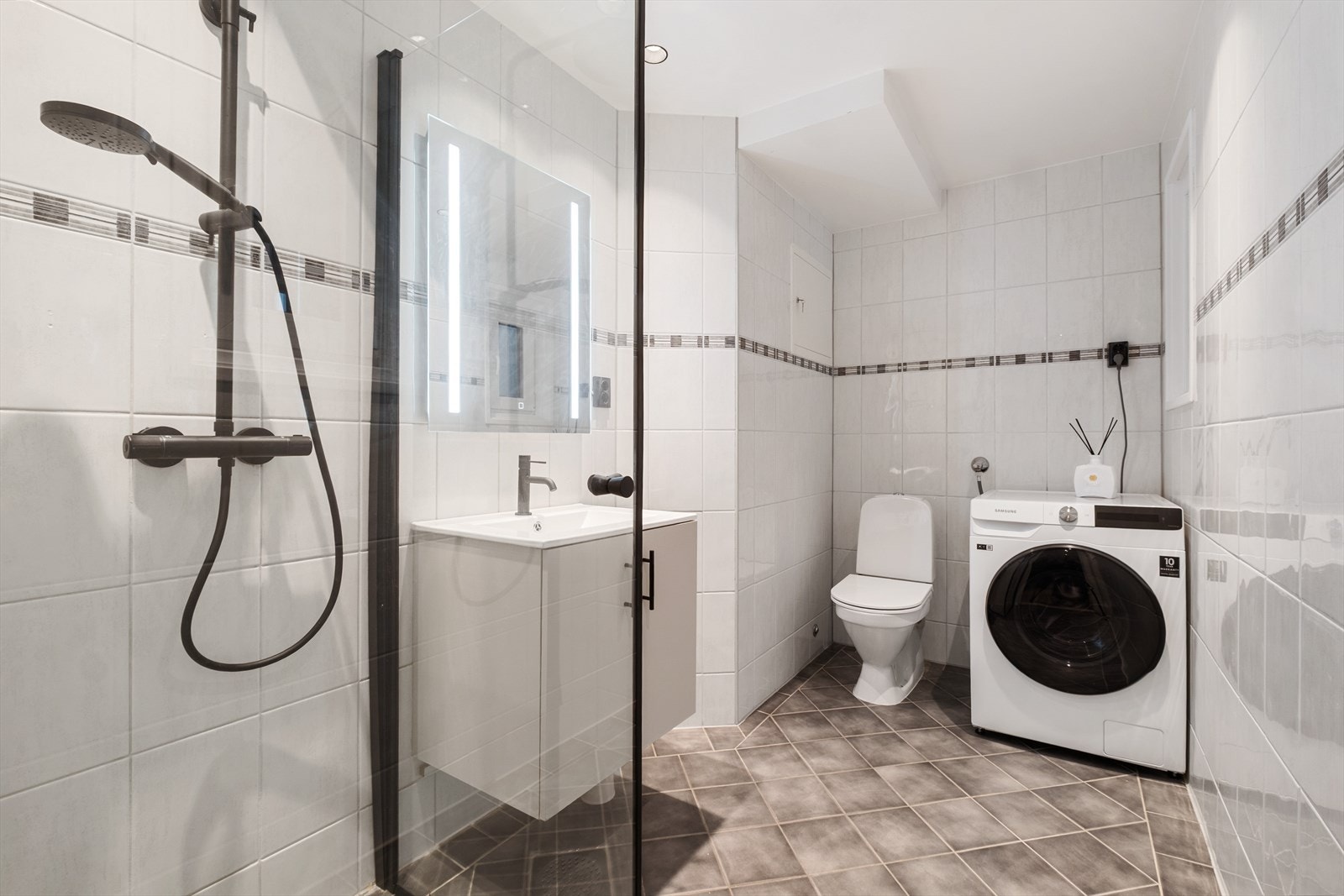 Innredet med servantskap, speil med integrert belysning, gulvmontert toalett, dusjhjørne med dusjvegger og opplegg for vaskemaskin. Galleribilde