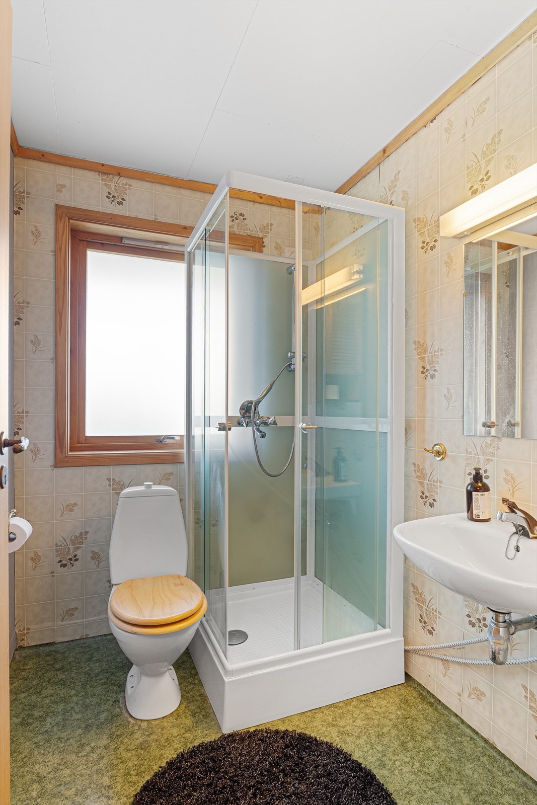 Bad trolig fra byggeår i underetasjen med belegg på gulv og tapet på vegg. Badet har toalett, dusjkabinett og vask. Galleribilde