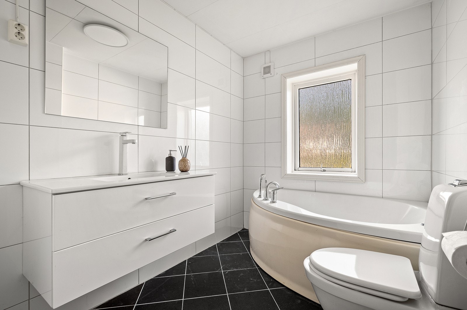 Bad i 1.etasje med fliser på vegg og gulvflater. Ventilasjon består av el-ventil i vegg og sluk i PVC/plast. Bad ble oppgradert på overflater en gang på 2000 tallet. Galleribilde