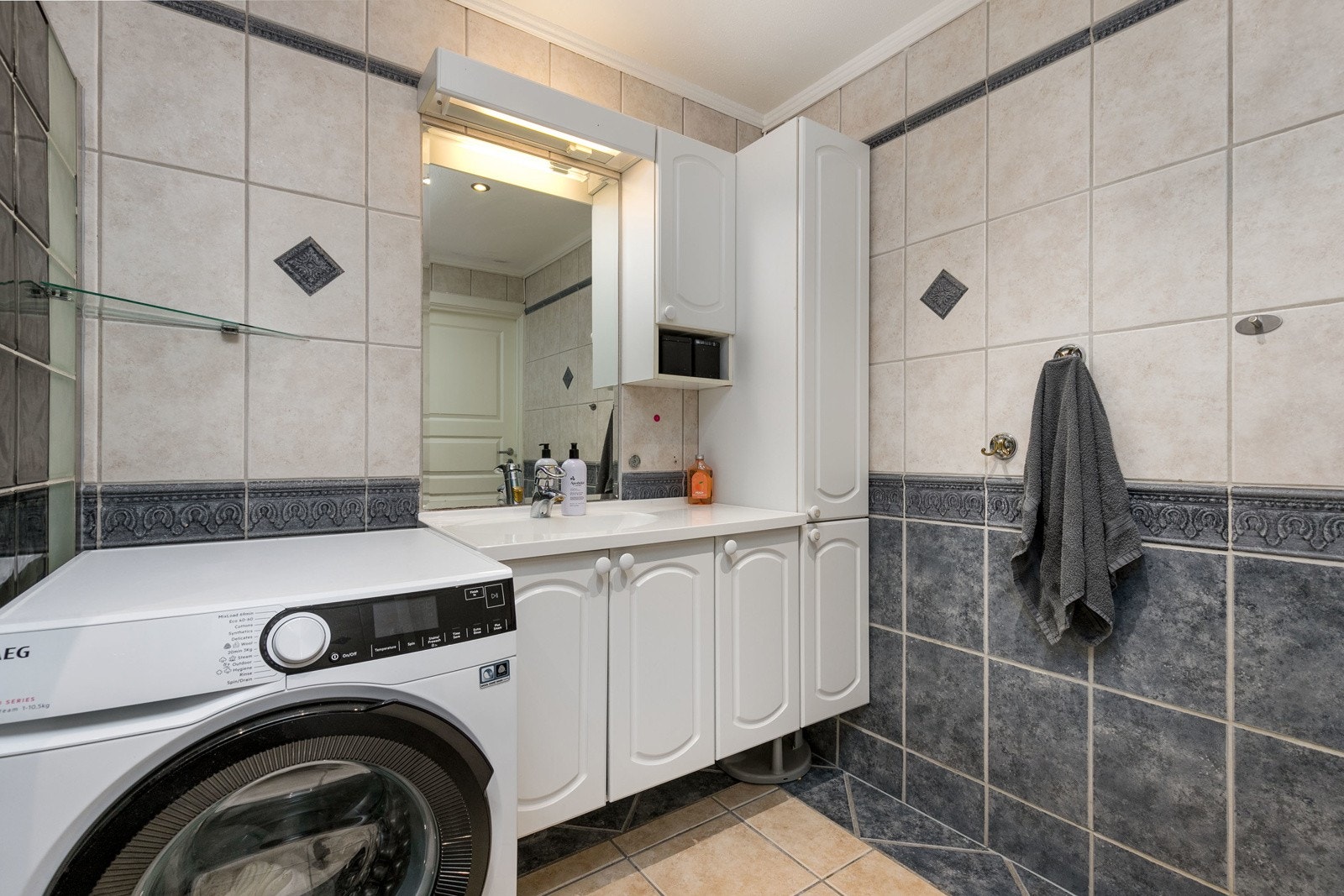 Badet er flislagt med varmekabler i gulvet og av eldre dato. Innredningen består av servantmøblement med under- og overskap, speil og frittstående toalett. Galleribilde