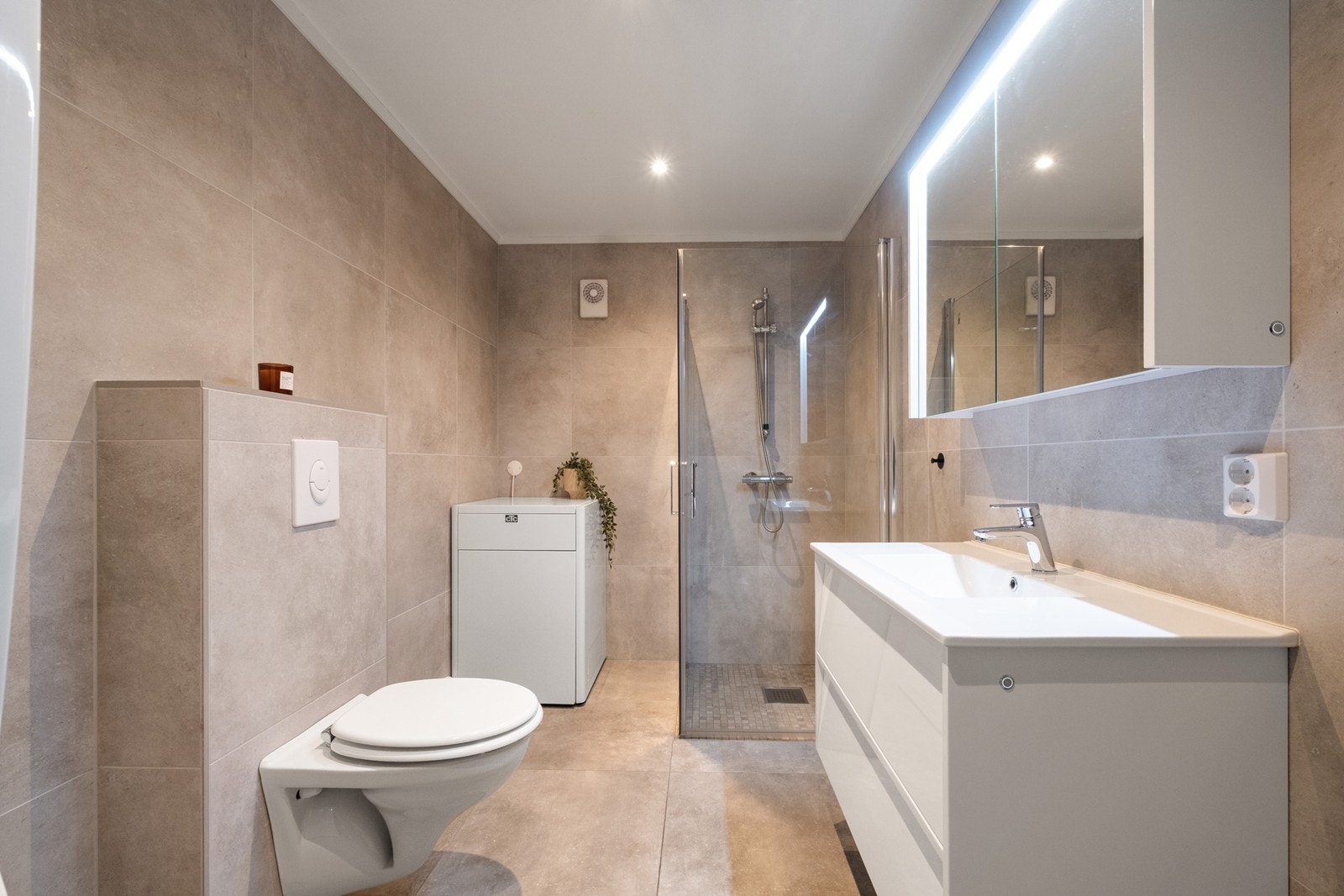 Flislagt bad utstyrt med vegghengt wc, dusjhjørne med glassvegger og heldekkende servant med både underskap og belyst speilskap. Galleribilde