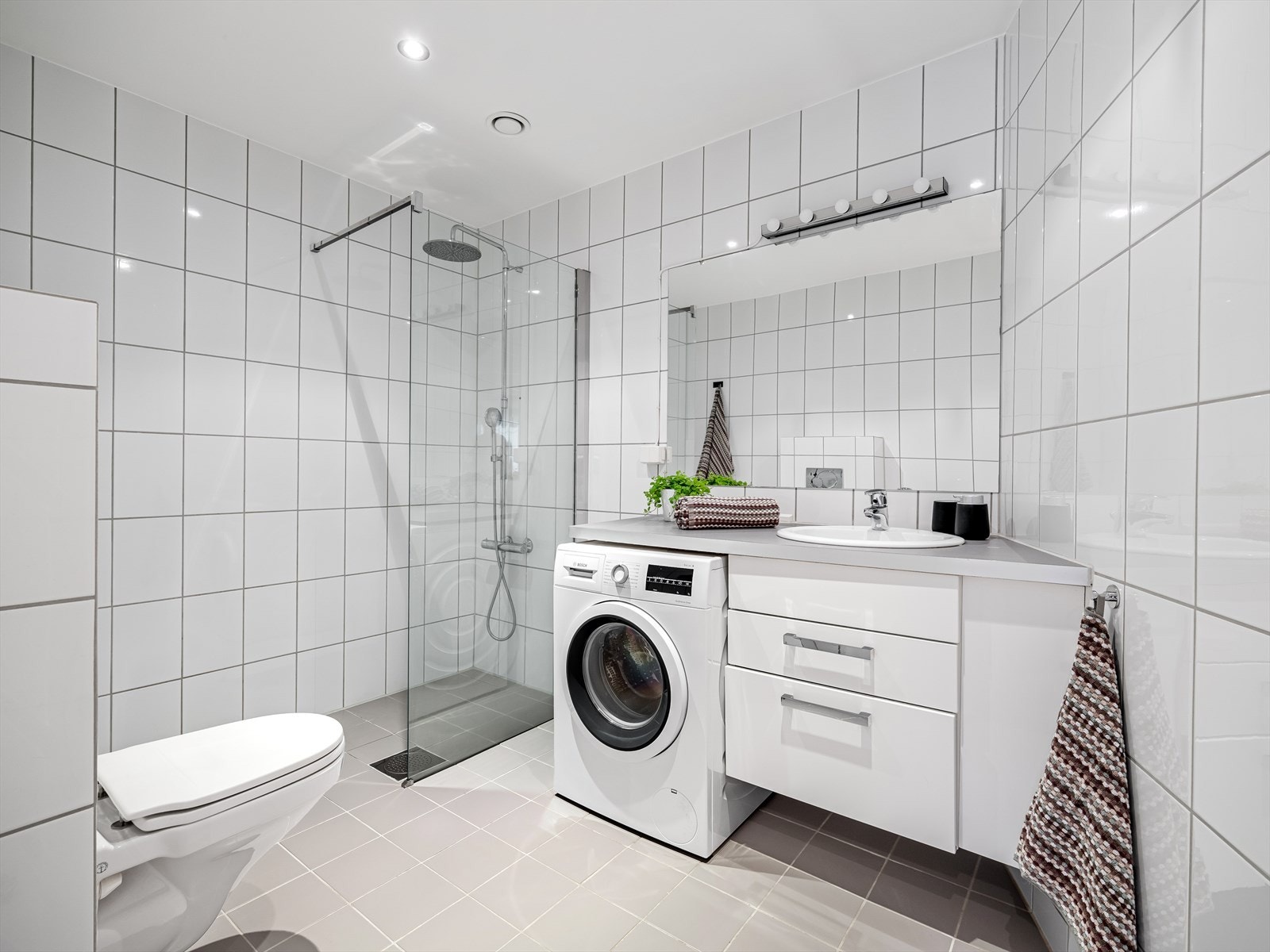 Badet er innredet med vegghengt toalett og dusjhjørne med glassvegg og regnfallsdusj. Skapinnredning med skuffer, benkeplate og nedfelt servant Galleribilde