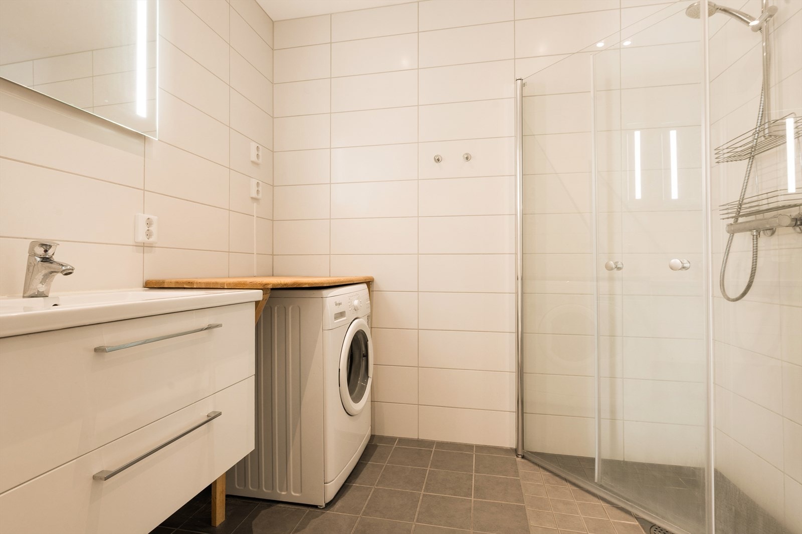 Badet er innredet med dusj på vegg og dusjdører i herdet glass, helservant på skuffeseksjon, speil med belysning og klosett. Galleribilde