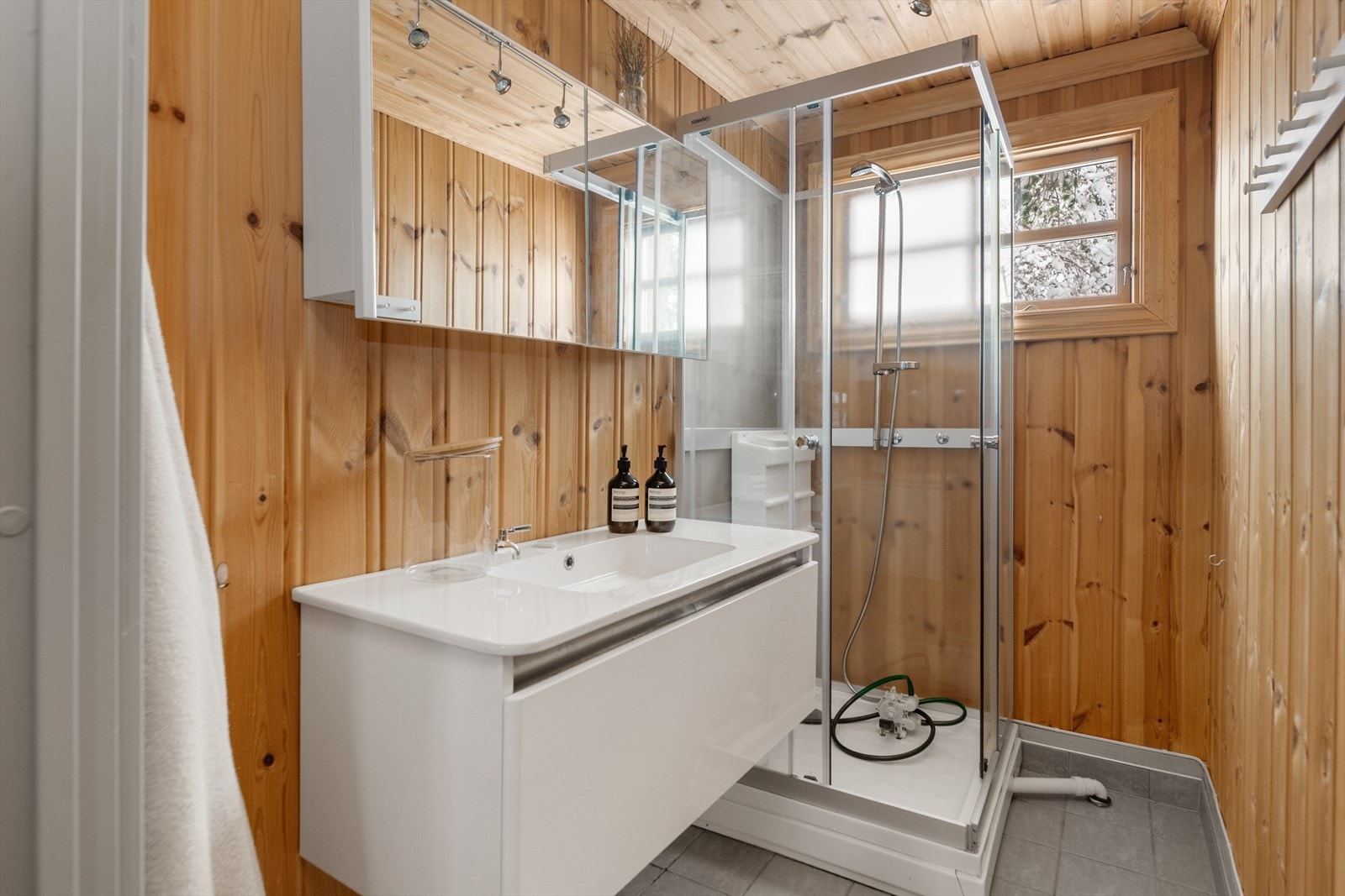 Baderommet har fliser på gulvet med gulvvarme, og beiset panel på vegger. Rommet innehar dusjkabinett med pumpe-dusj og baderomsinnredning med skuff og skap med speildører. Galleribilde