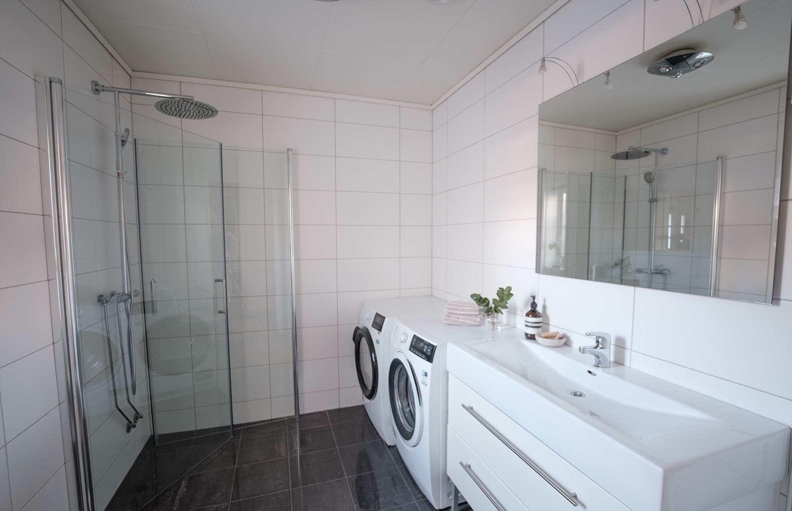 Badet i første etasje har fliser på gulv og vegger. Her er dusjdører som kan slås inn, innredning med servant, toalett og opplegg for vaskemaskin og tørketrommel. Galleribilde