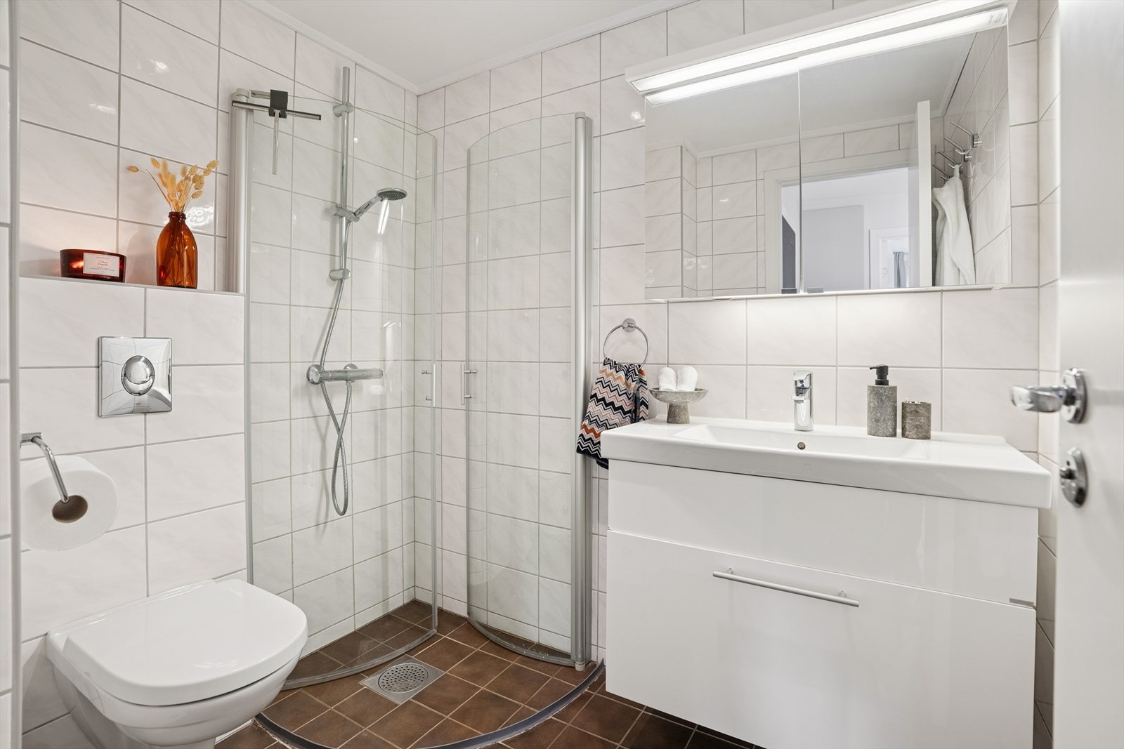 Baderommet er helfliset og moderne, med vegghengt toalett, servant med praktisk innredning og et effektivt dusjhjørne med innfellbare glassdører. Et stilrent rom med gode materialvalg. Galleribilde