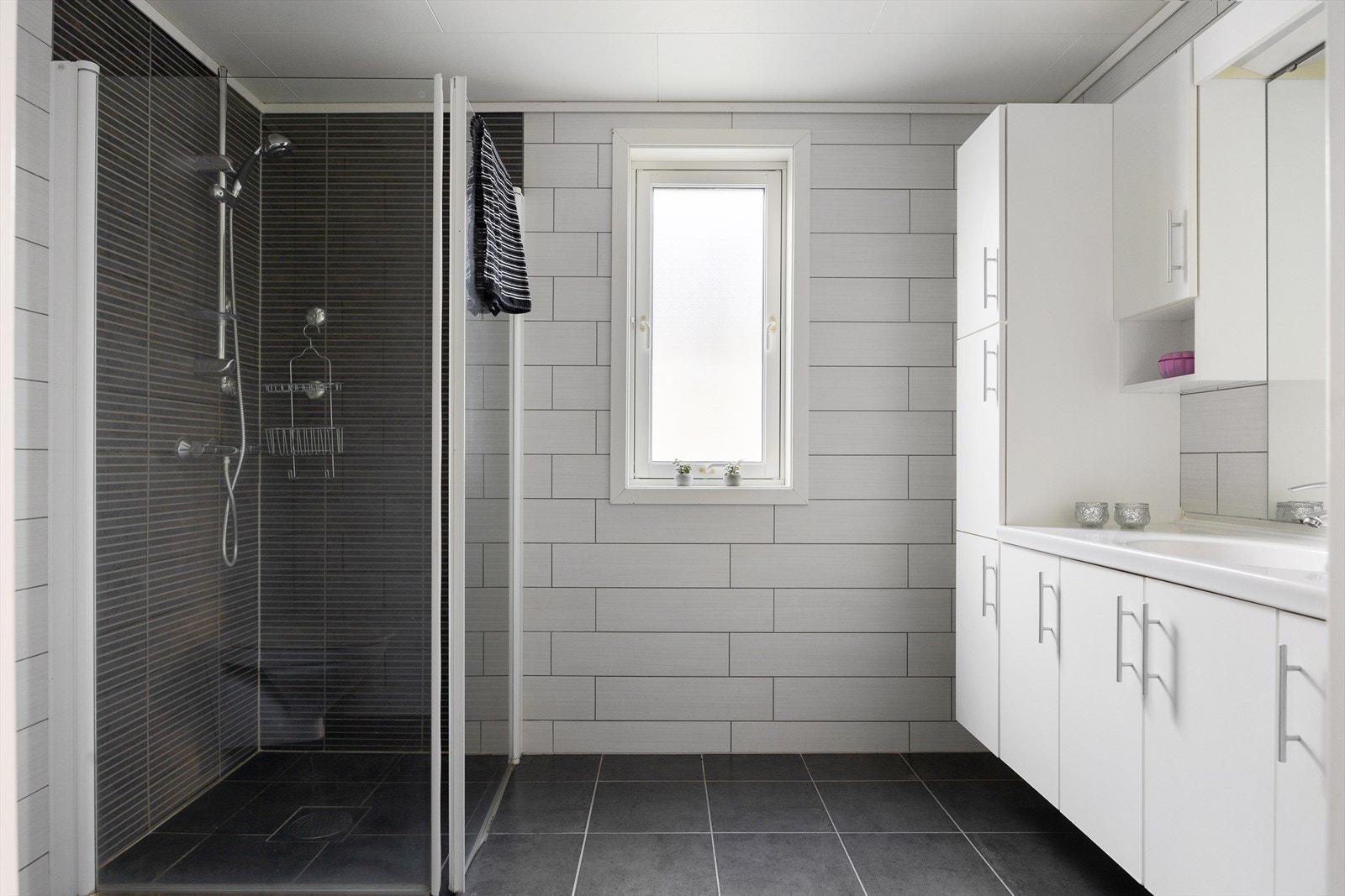 Badet er innredet med servant nedfelt i benkeskap, veggskap for praktisk oppbevaring, speil, et moderne vegghengt toalett og dusjhjørne med glassdører. Galleribilde