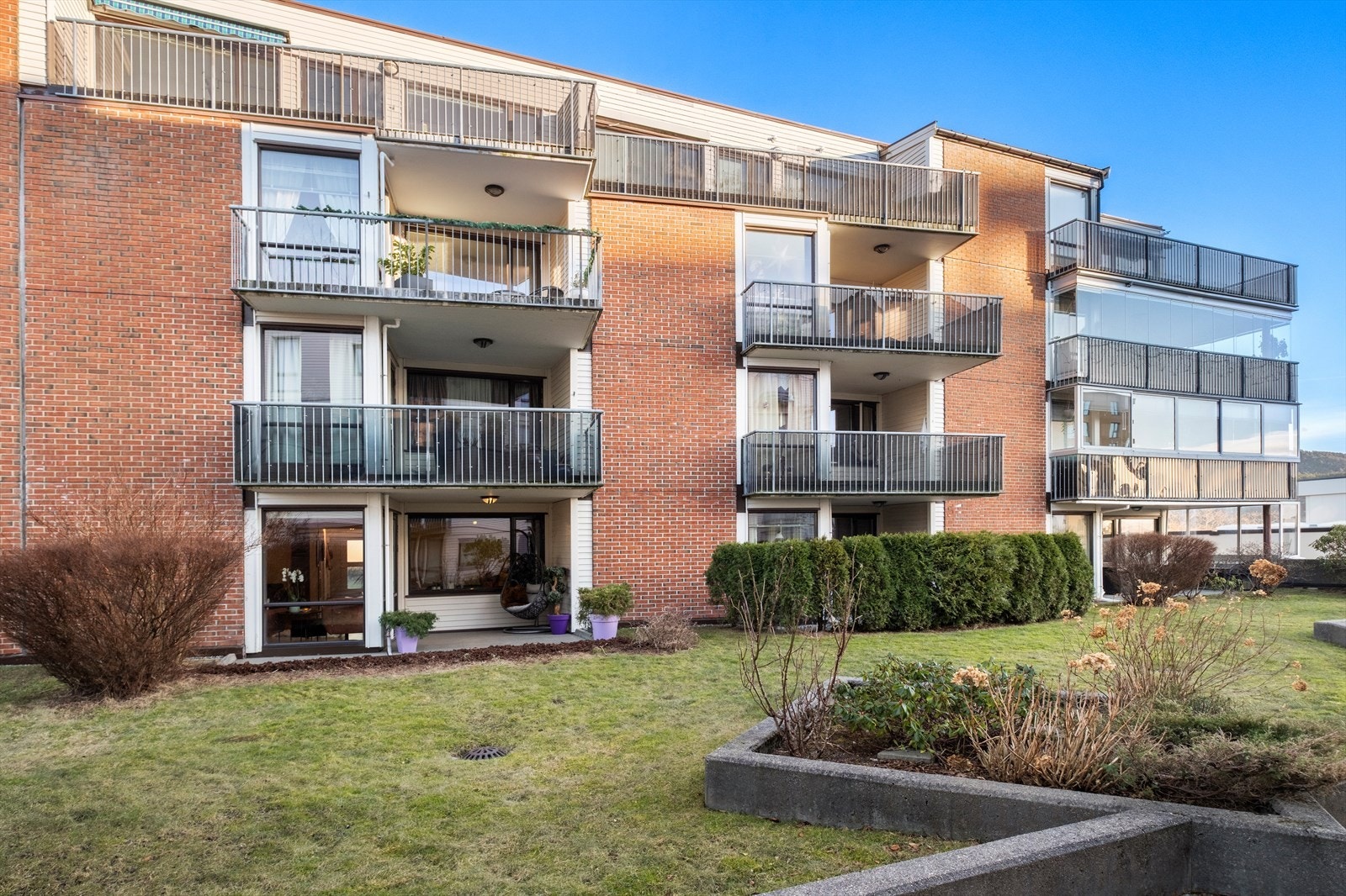 Gressplen og beplantning omgir bygget, som har teglsteinfasade og balkonger. Galleribilde