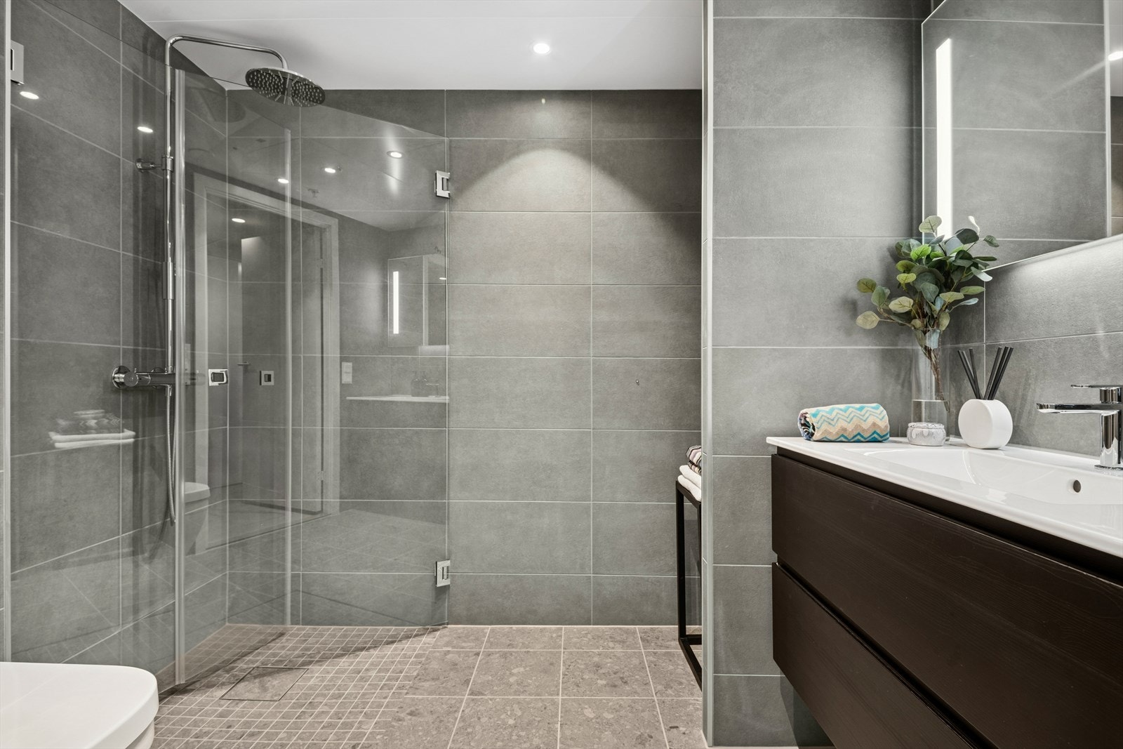 Badet er innredet med et dusjhjørne med innfellbare glassdører og regnfallsdusj, veggmontert toalett, moderne innredning med heldekkende servant og speil med belysning. Galleribilde