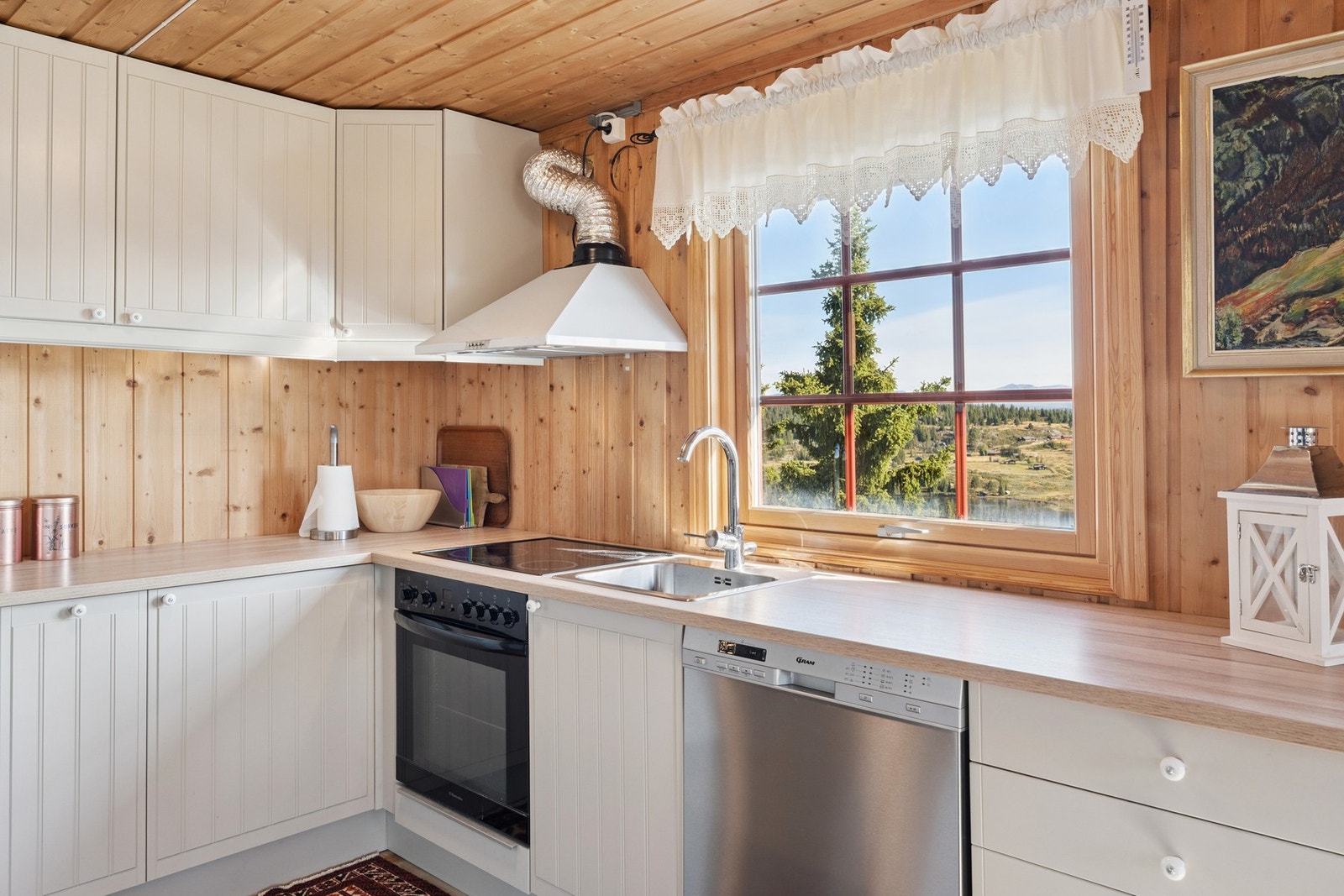 Lysmalt kjøkkeninnredning med oppvaskkum nedfelt i laminat benkeplate. Integrerte hvitevarer som komfyr og koketopp, frittstående oppvaskmaskin og kjøl-/fryseskap. Galleribilde