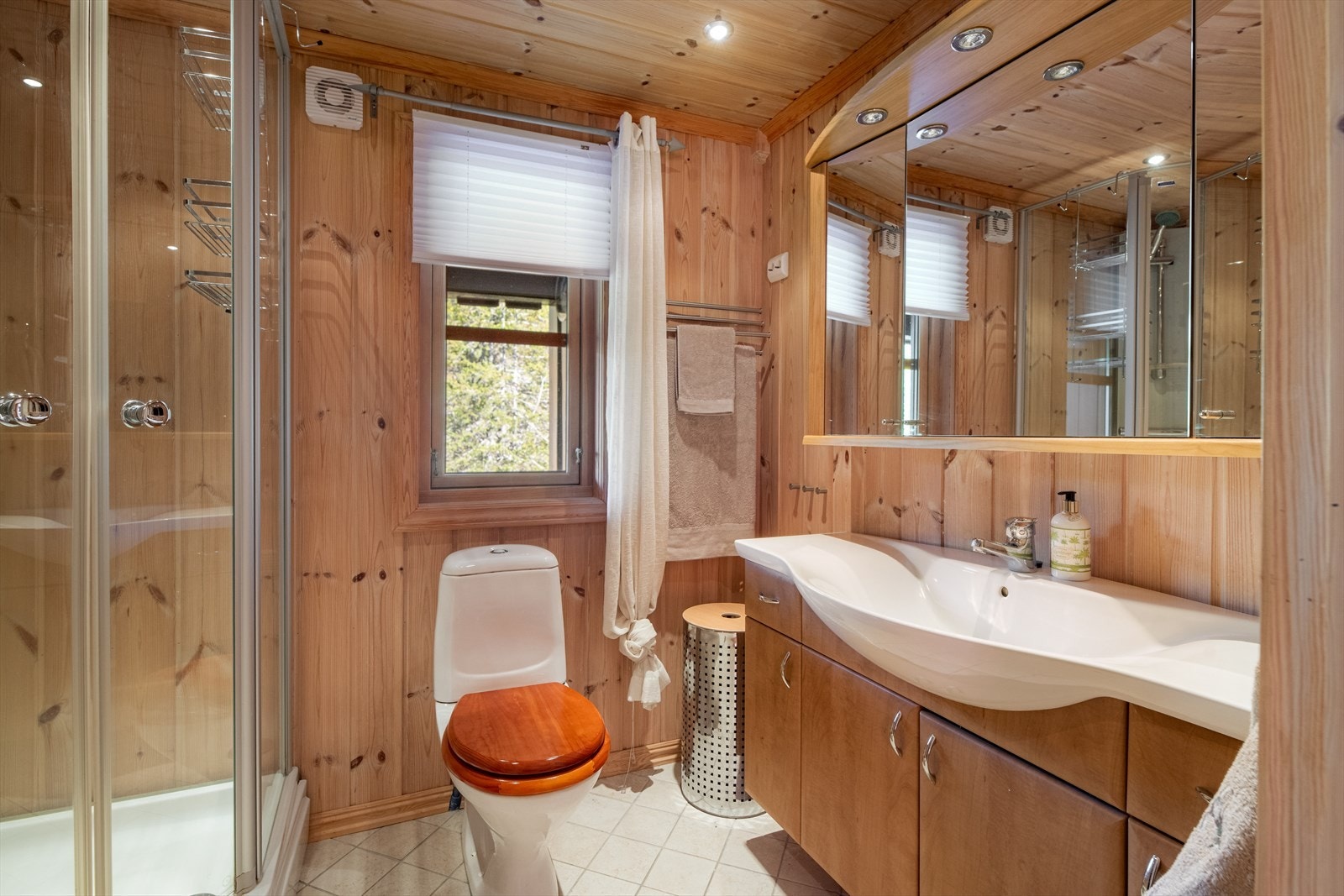 Pent baderom som inneholder heldekkende servant med underskap, speilskap med lys, gulvmontert toalett og dusjkabinett. Galleribilde