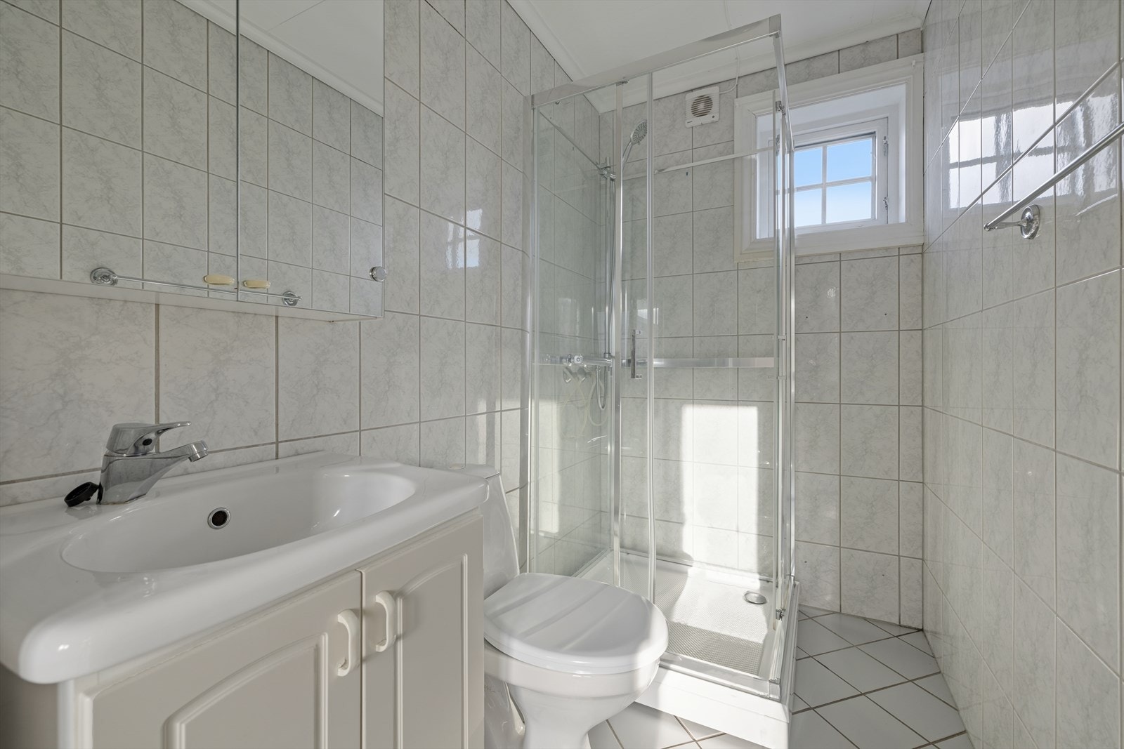 Badet er av eldre alder og har en enkel innredning med lyse fliser, varmekabler, nedfelt servant, gulvfestet toalett og dusjkabinett. Galleribilde