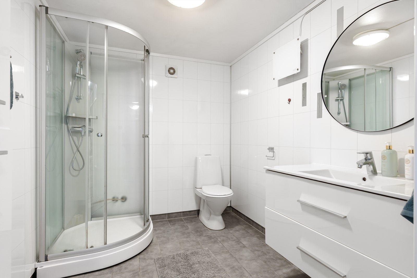 Tidløst bad med moderne baderomsinnredning bestående av benkeskap og høyskap med glatte fronter, speil over servant, dusjkabinett, servant og toalett Galleribilde