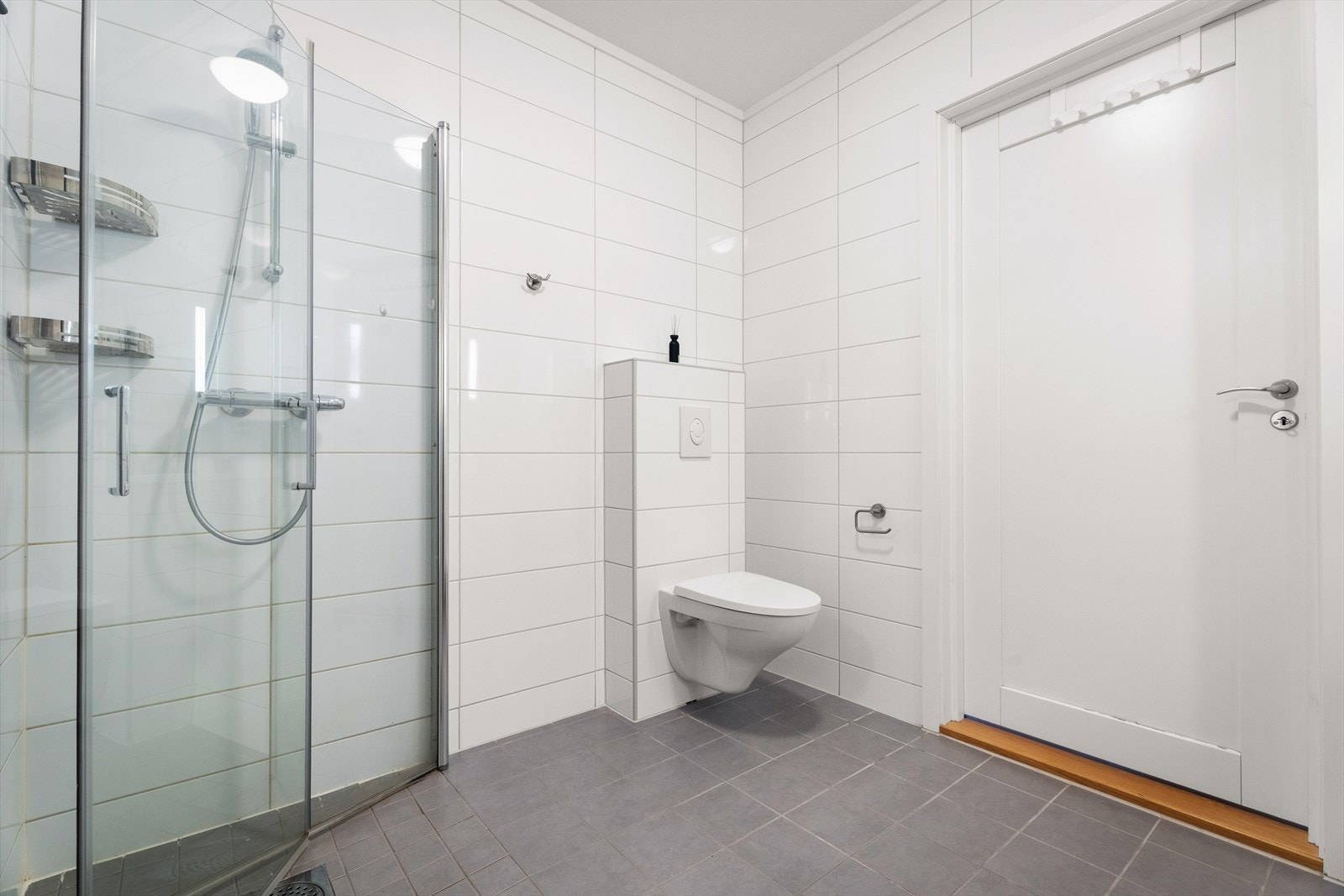 Badet inneholder dusjhjørne med innfellbare glassdører og et vegghengt toalett. Galleribilde