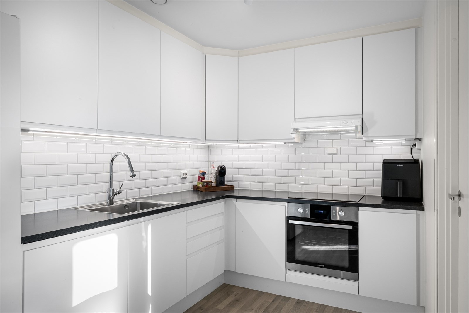 Flott kjøkken med pen kjøkkeninnredning i hvit utførelse som inneholder integrert komfyr, platetopp, oppvaskamskin og vifte. Galleribilde