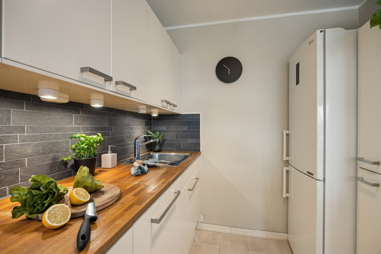 Innredning med slette lakkerte fronter, heltre benkeplate med fliser på bakvegg, og innfelte hvitevarer bestående av stekeovn, induksjonsplatetopp og oppvaskmaskin samt et frittstående kombinert kjøle- og fryseskap. Galleribilde