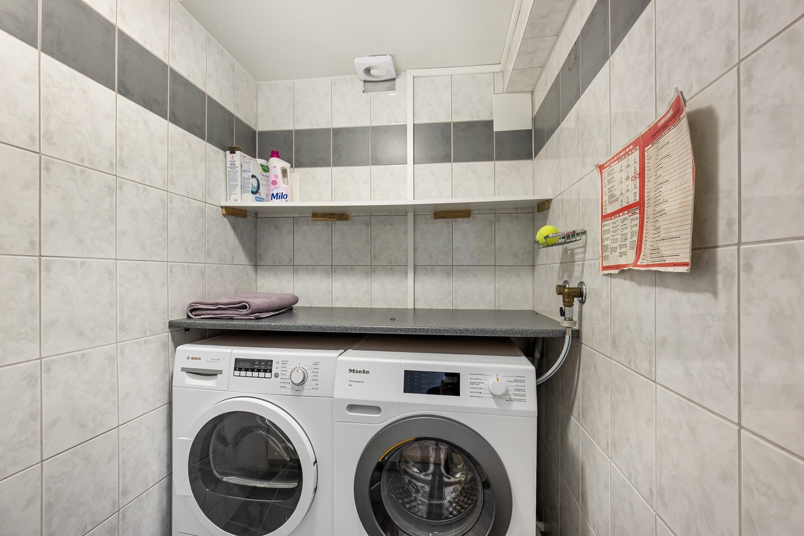 Flislagt vaskerom utstyrt med benkeinnredning med opplegg for vaskemaskin og tørketrommel under benk. Elektronisk styrt vifte fra 2024 Galleribilde