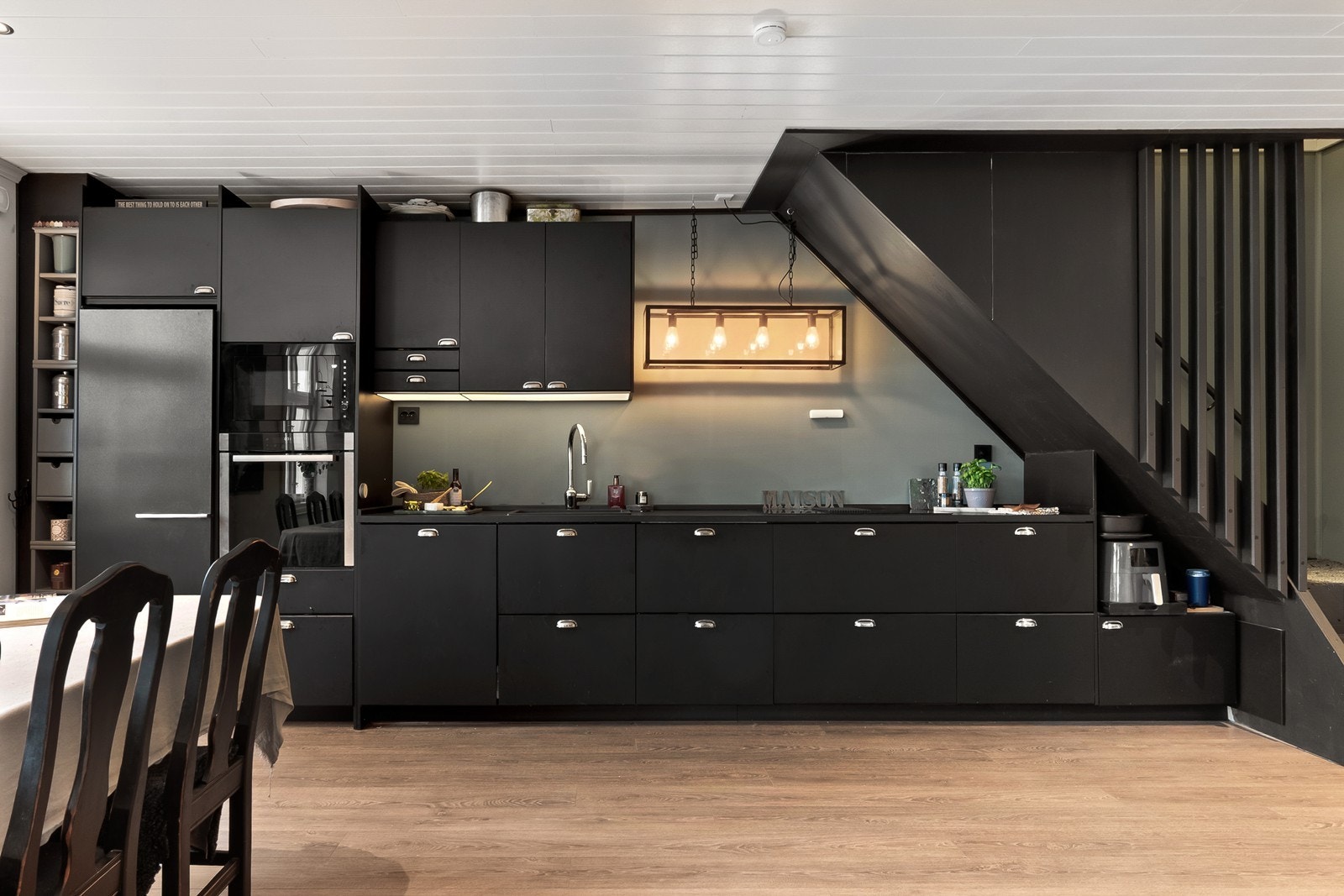 Løsningen er kompakt langs hele veggen med gode arbeidsflater og oppbevaringsmuligheter. Integrerte hvitevarer består platetopp med ventilator, stekeovn, mikroovn og oppvaskmaskin, samt frittstående kjøl-/fryseskap. Galleribilde