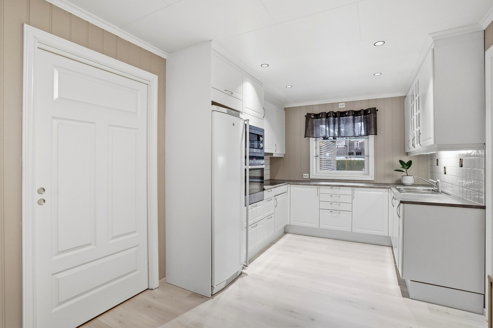 Pent kjøkken med innredning fra Sigdal som har hvite fronter, benkeplate i laminat og integrerte hvitevarer som stekeovn, mikroovn, platetopp og oppvaskmaskin, samt frittstående kjøleskap. Galleribilde