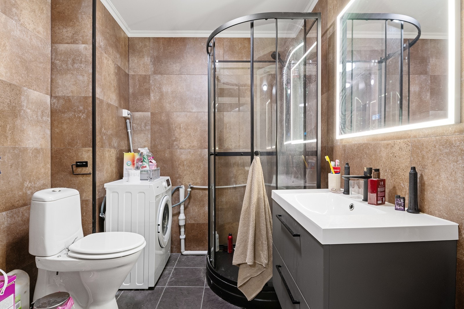 Badet/vaskerom i 1. etasje har baderomsplater på vegg og vinylbelegg på gulv. Det er utstyrt med toalett, nedfelt vask i skuffeseksjon, dusjkabinett og uttak for vaskemaskin. Galleribilde