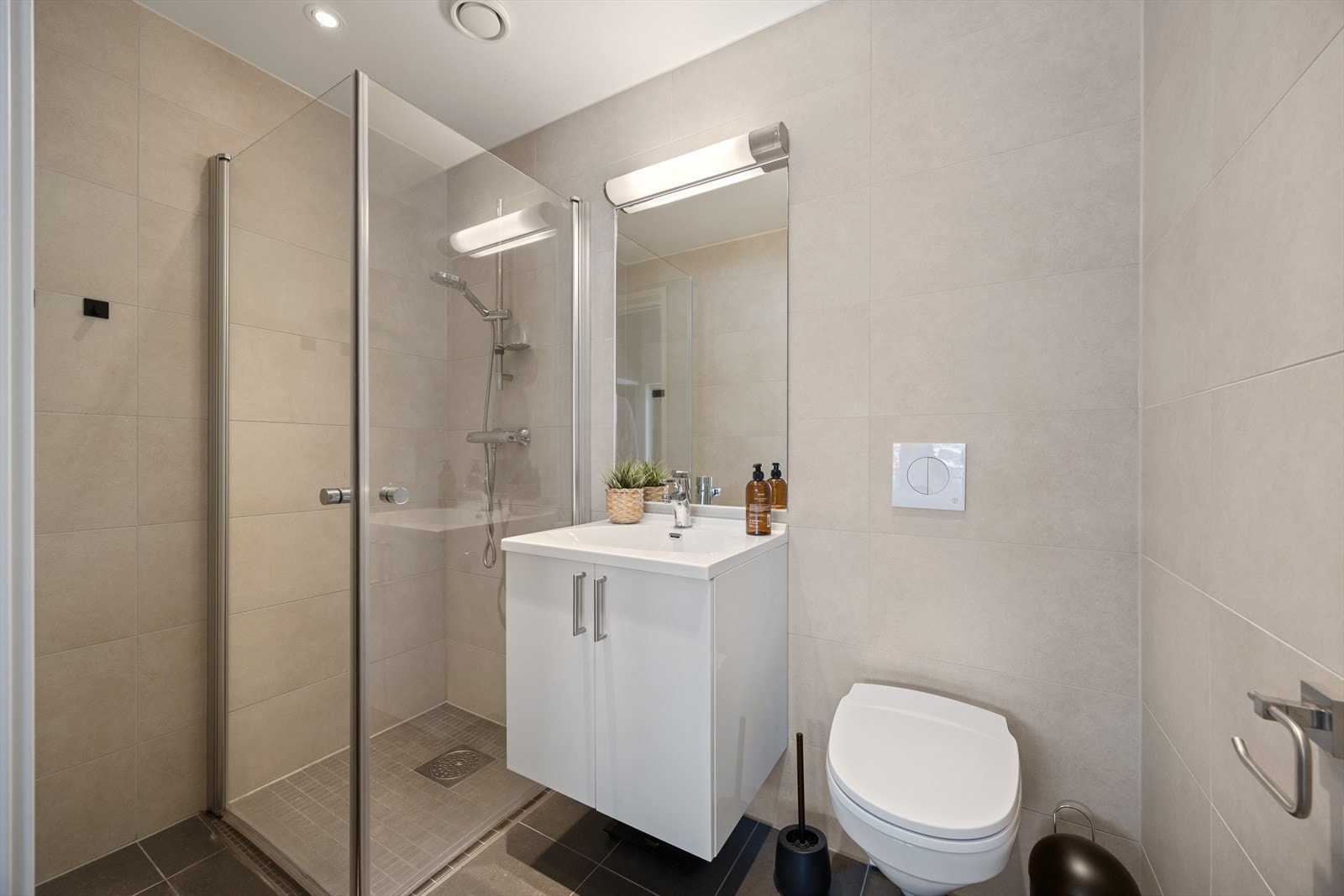 Badet er pent innredet. Tidløse grå fliser på gulvet og beige fliser på vegg. Varmekabler på gulvet. Badet er ellers utstyrt med dusjhjørne, vegghengt toalett og baderomsinnredning med nedfelt servant. Galleribilde