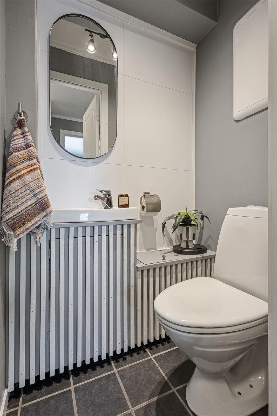 Toalettrommet er innredet med grå fliser på gulvet og en kombinasjon av baderomsplater og malte veggflater. Rommet er ellers utstyrt med toalett og innredning med servant. Galleribilde