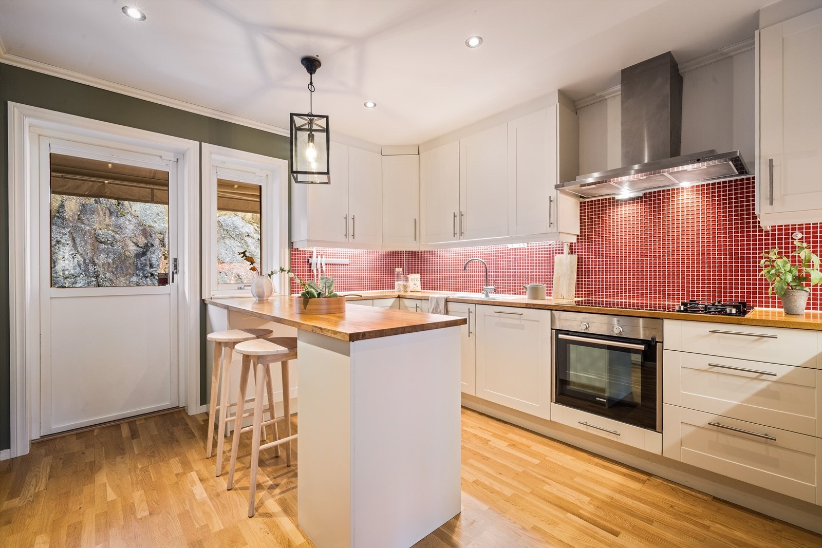 Pent kjøkken i hvit utførelse med profilerte fronter, heltre benkeplate og fliser på vegg over benkeplate. De store vindusflatene gir godt med naturlig lys. Galleribilde