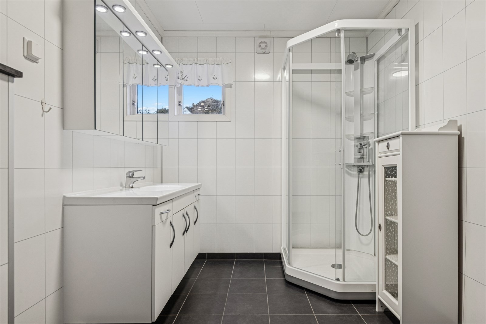 Baderommet har flislagt gulv med elektriske varmekabler og er innredet med dusjkabinett samt servant med praktisk skapinnredning. Galleribilde