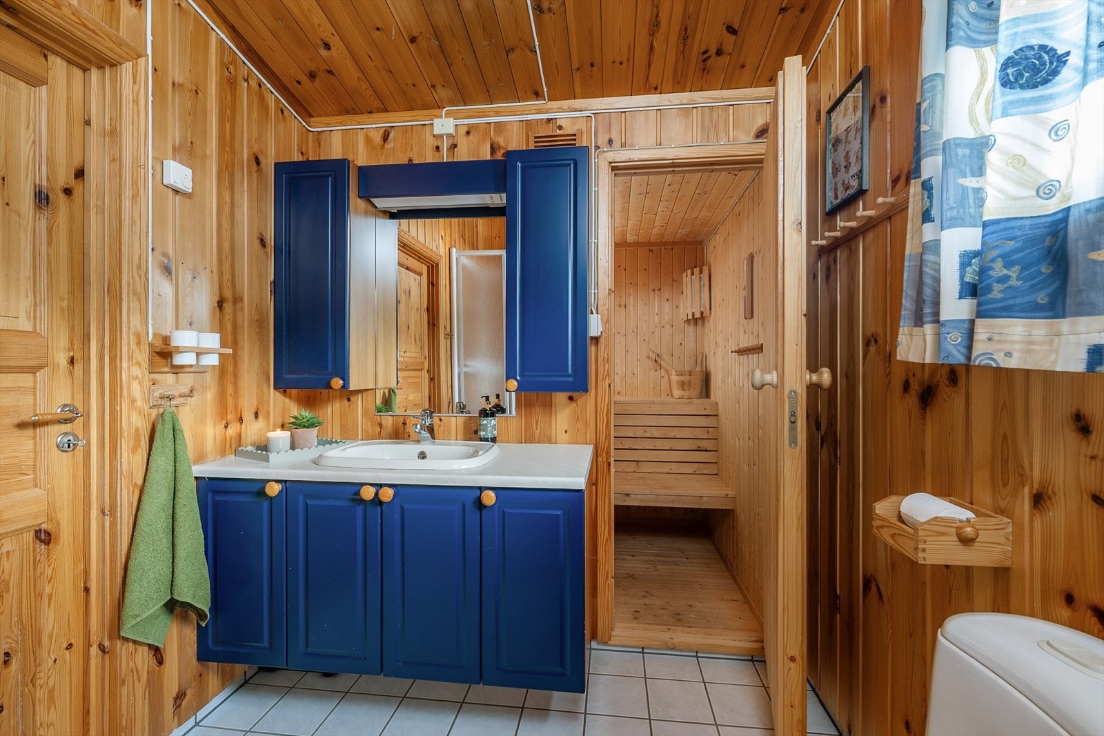 Praktisk bad med badstue. Baderomsinnredning med blåmalte fronter. Panelte tak og vegger. Fliser på gulv. Galleribilde