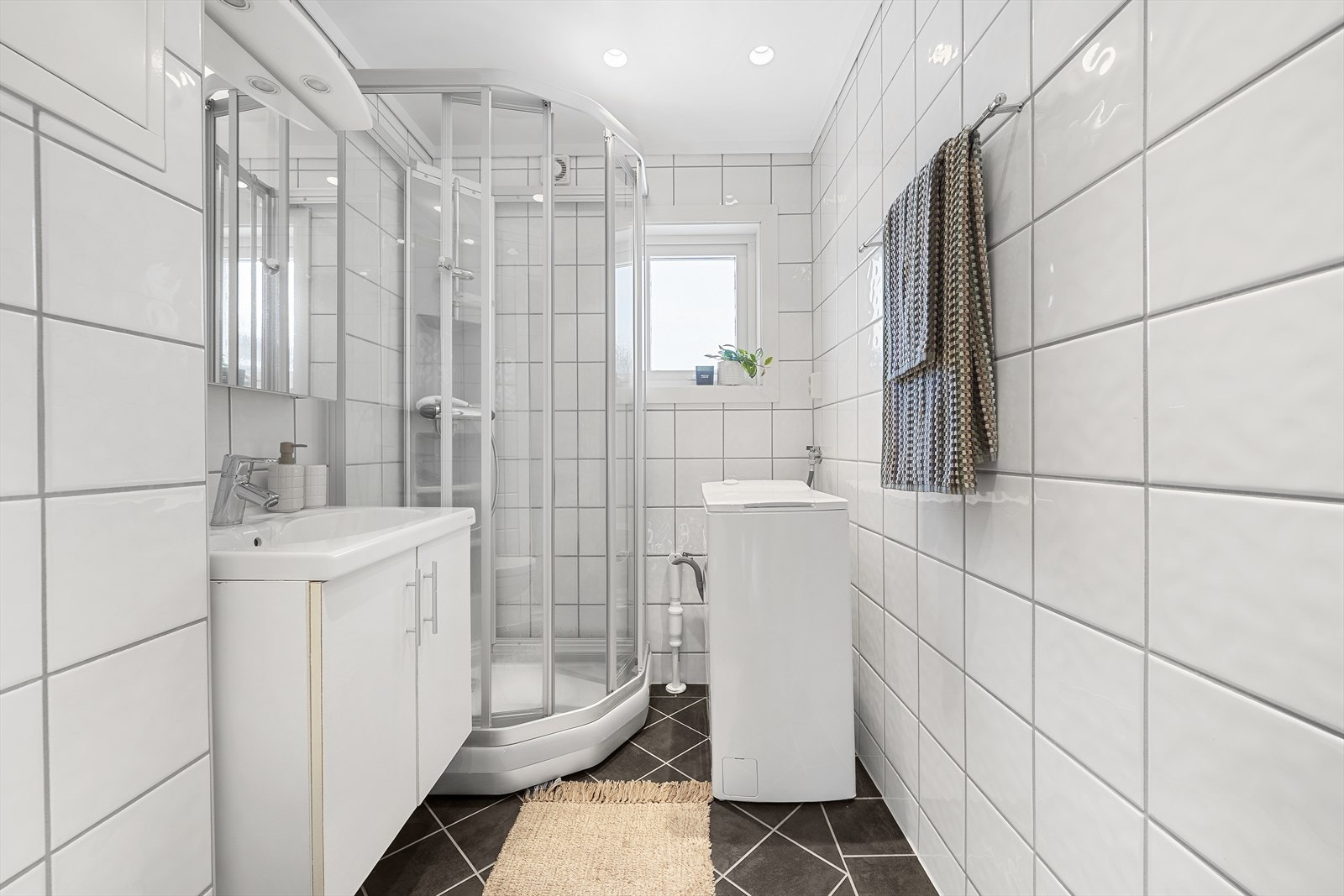 Pent flislagt baderom med varmekabler i gulv. Rommet er utstyrt med servant over skapinnredning, speil, vegghengt toalett, dusjkabinett og opplegg til vaskemaskin Galleribilde