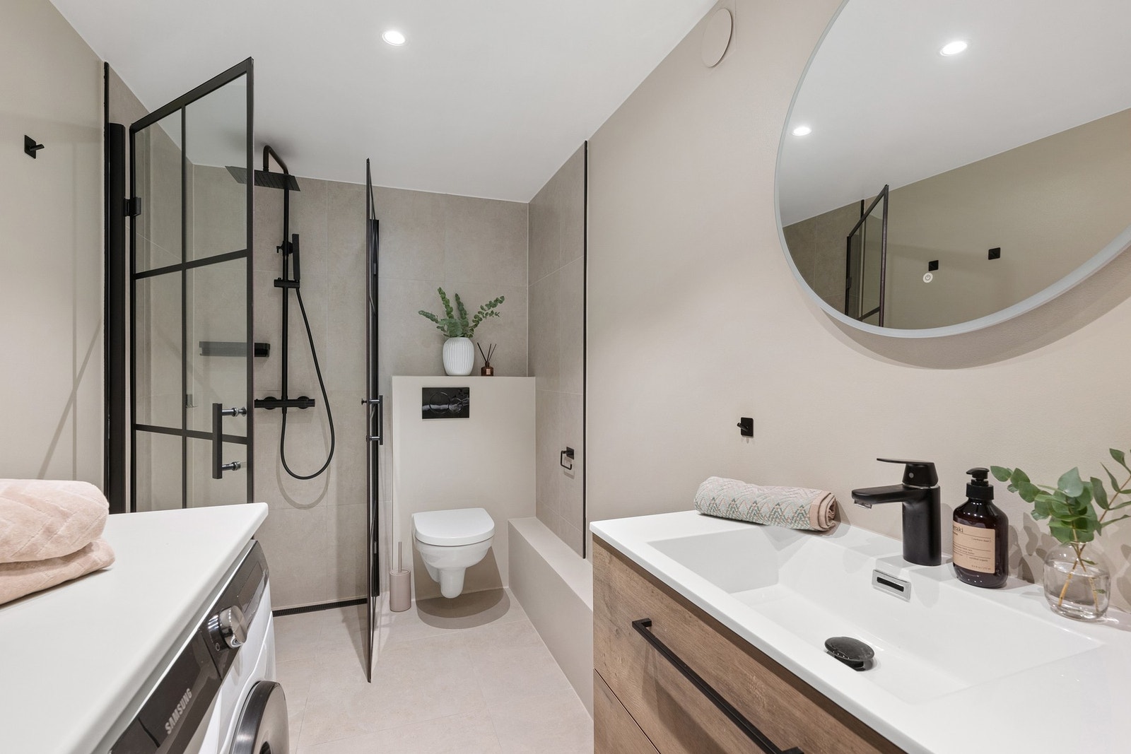 Moderne og råflott hovedbad med stilren innredning, rundt speil med integrert belysning og sorte detaljer. Dagens eiere har i tillegg montert baderomsskap og praktisk arbeidsbenk over vaskemaskin. Galleribilde